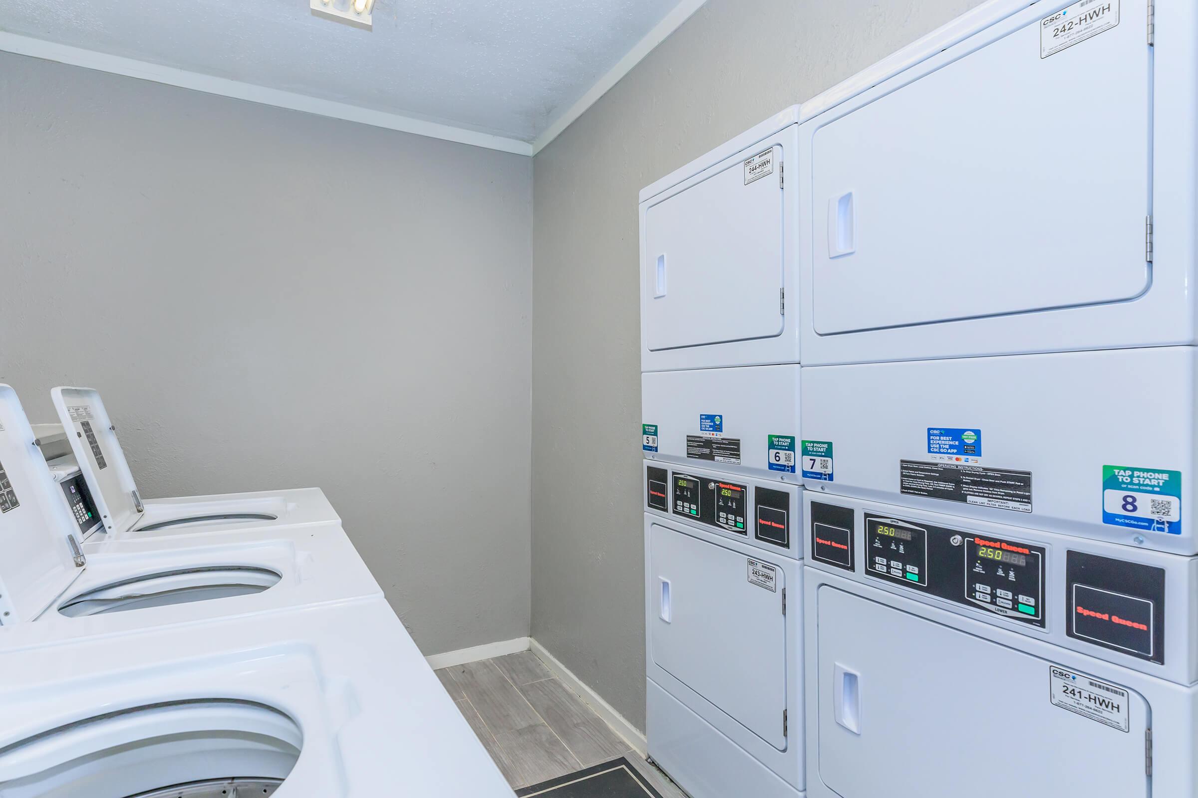 A clean and modern laundry room featuring several stacked washing machines and dryers. The walls are painted light gray, and the flooring is light-colored. The machines have clear indicators and labels for operation, providing a tidy and functional space for doing laundry.