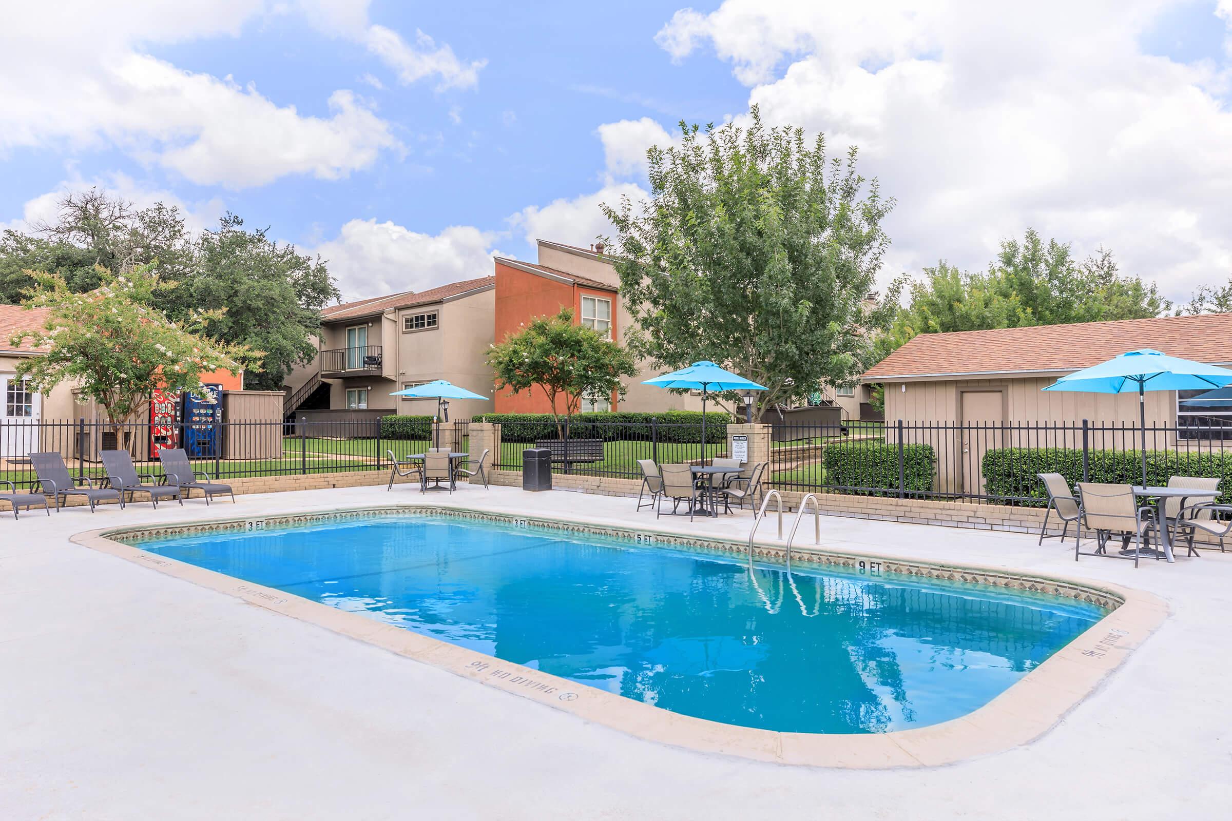 A clear blue swimming pool surrounded by lounge chairs and tables with umbrellas. The area is bordered by green trees and shrubs, with two buildings visible in the background, adding a residential ambiance. The sky is partly cloudy, suggesting a pleasant day.