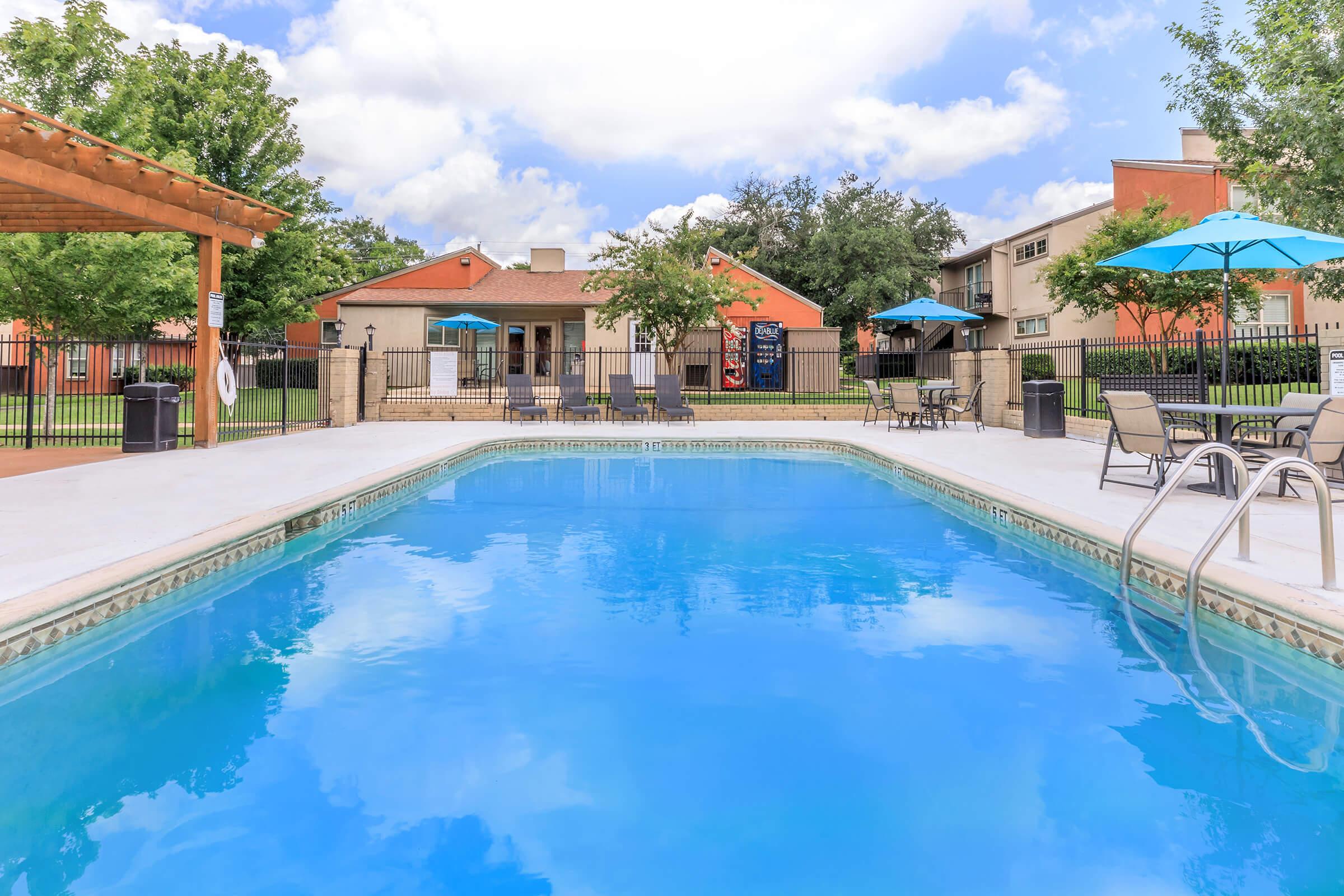 A clear blue swimming pool surrounded by lounge chairs and tables with umbrellas, set in a well-maintained outdoor area. In the background, there are buildings with green lawns and trees, providing a relaxing atmosphere. The sky is partly cloudy, adding to the inviting environment.
