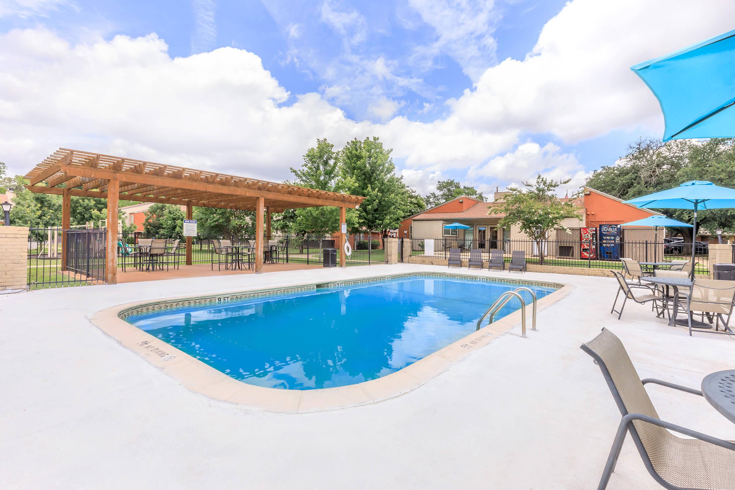 A clear swimming pool surrounded by a wooden pergola, lounge chairs, and patio tables. Lush green trees and a few buildings are visible in the background, along with blue umbrellas offering shade. The sky is partly cloudy, creating a tranquil atmosphere.