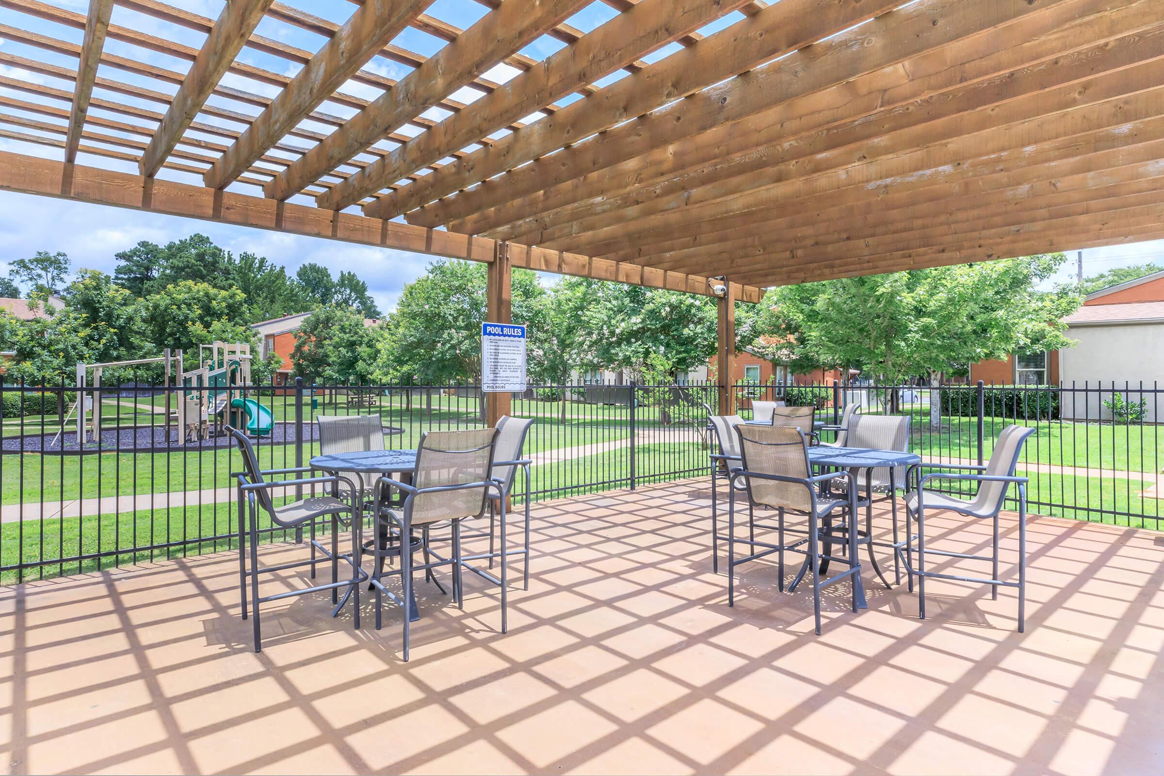 Outdoor seating area under a wooden pergola, featuring several tables and chairs. In the background, there is a playground surrounded by green grass and trees. The area is well-lit with a clear blue sky above. A pool rules sign can be seen on a nearby fence.