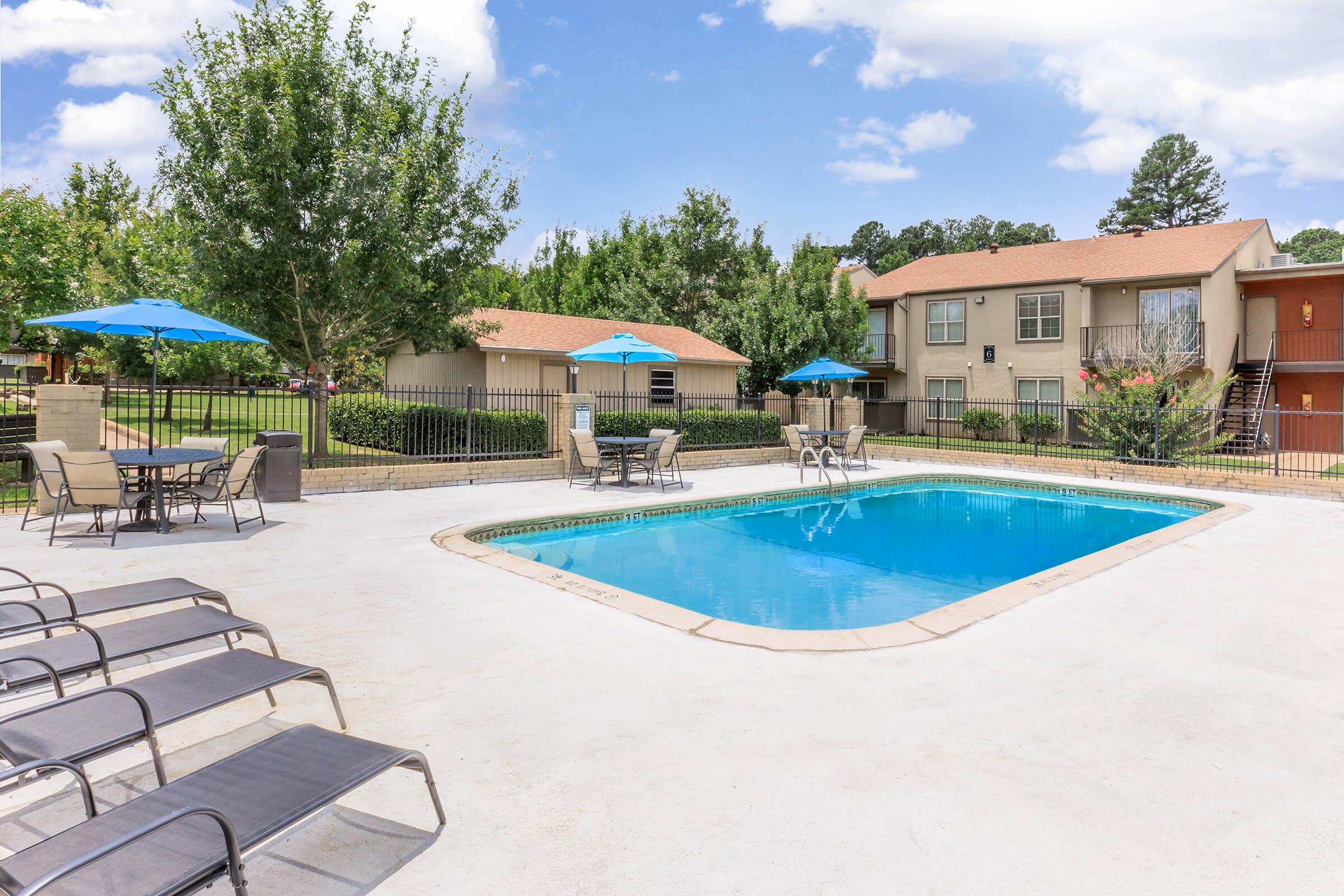 A clear blue swimming pool surrounded by lounge chairs and patio furniture under blue umbrellas. In the background, there are two multi-unit residential buildings and lush green trees under a partly cloudy sky. The area appears well-maintained and inviting for relaxation and leisure.
