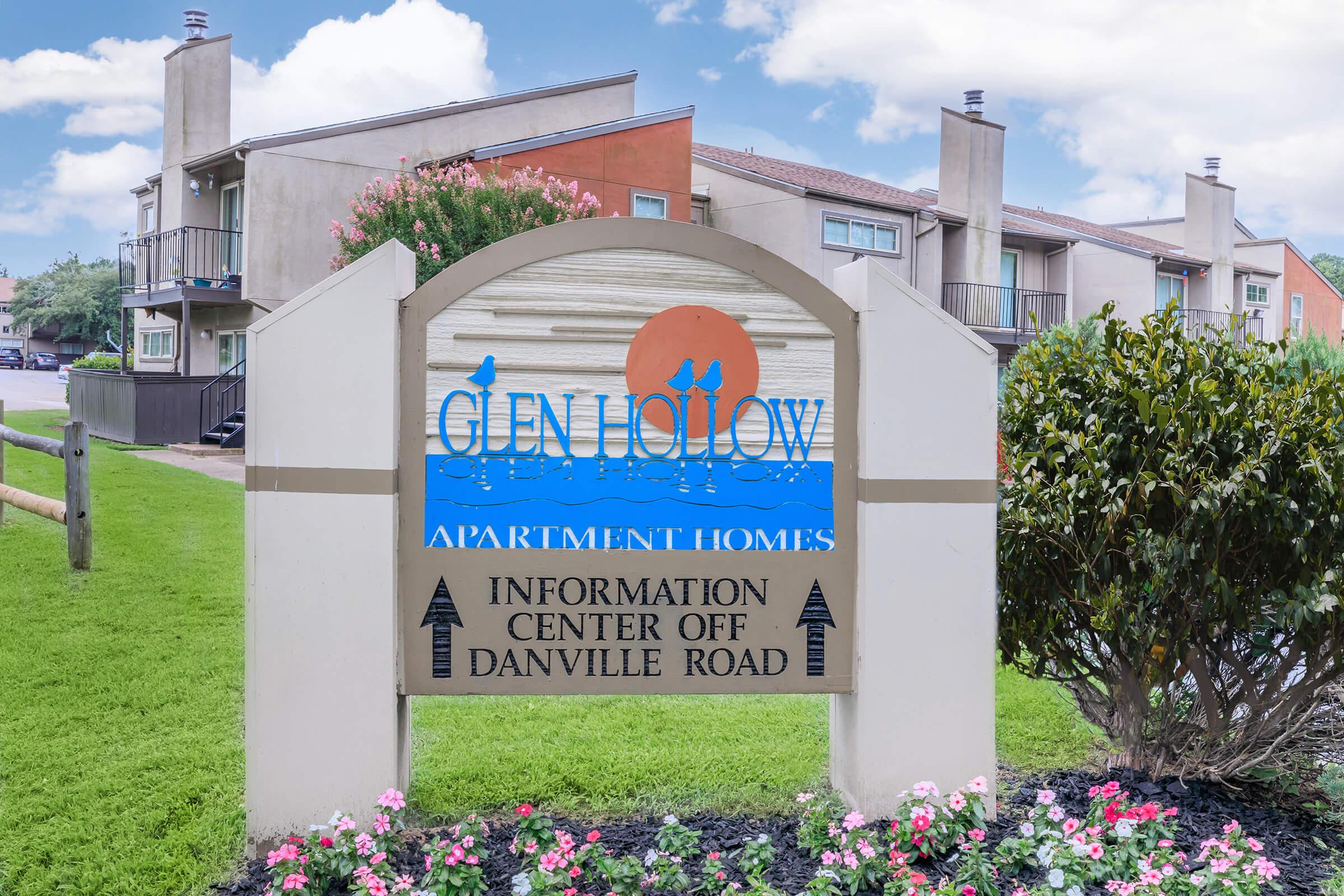 Sign for Glen Hollow Apartment Homes featuring a colorful logo with birds and a sun, surrounded by a landscaped area with flowers. The sign indicates that the information center is located off Danville Road. In the background, there are apartment buildings under a partly cloudy sky.