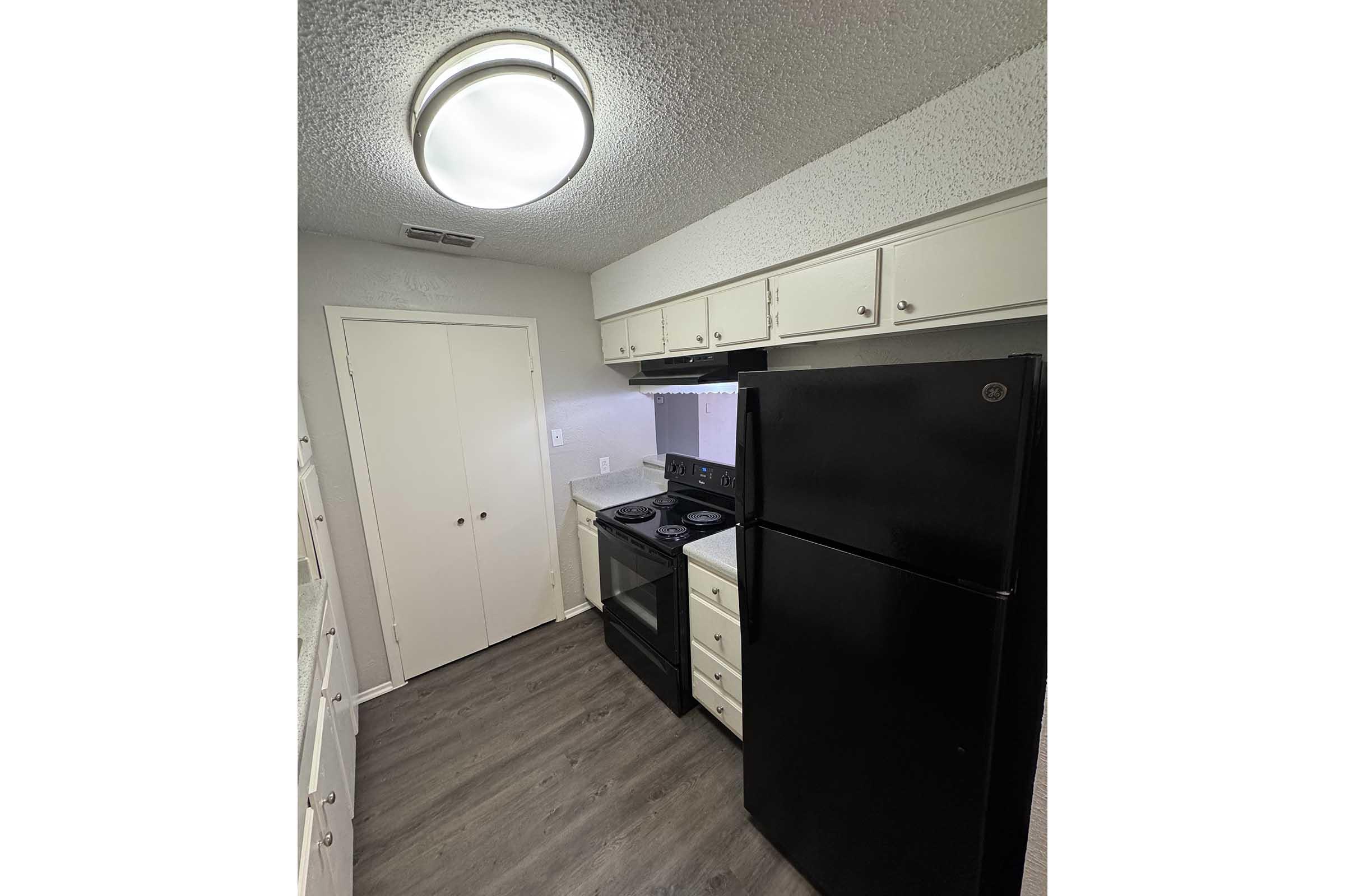 A compact kitchen featuring a black refrigerator and a black stove with an overhead microwave. The cabinets are white, and there is overhead lighting. The flooring is a light wood-like material, giving the kitchen a modern appearance. The layout is efficient, with a neutral color palette.