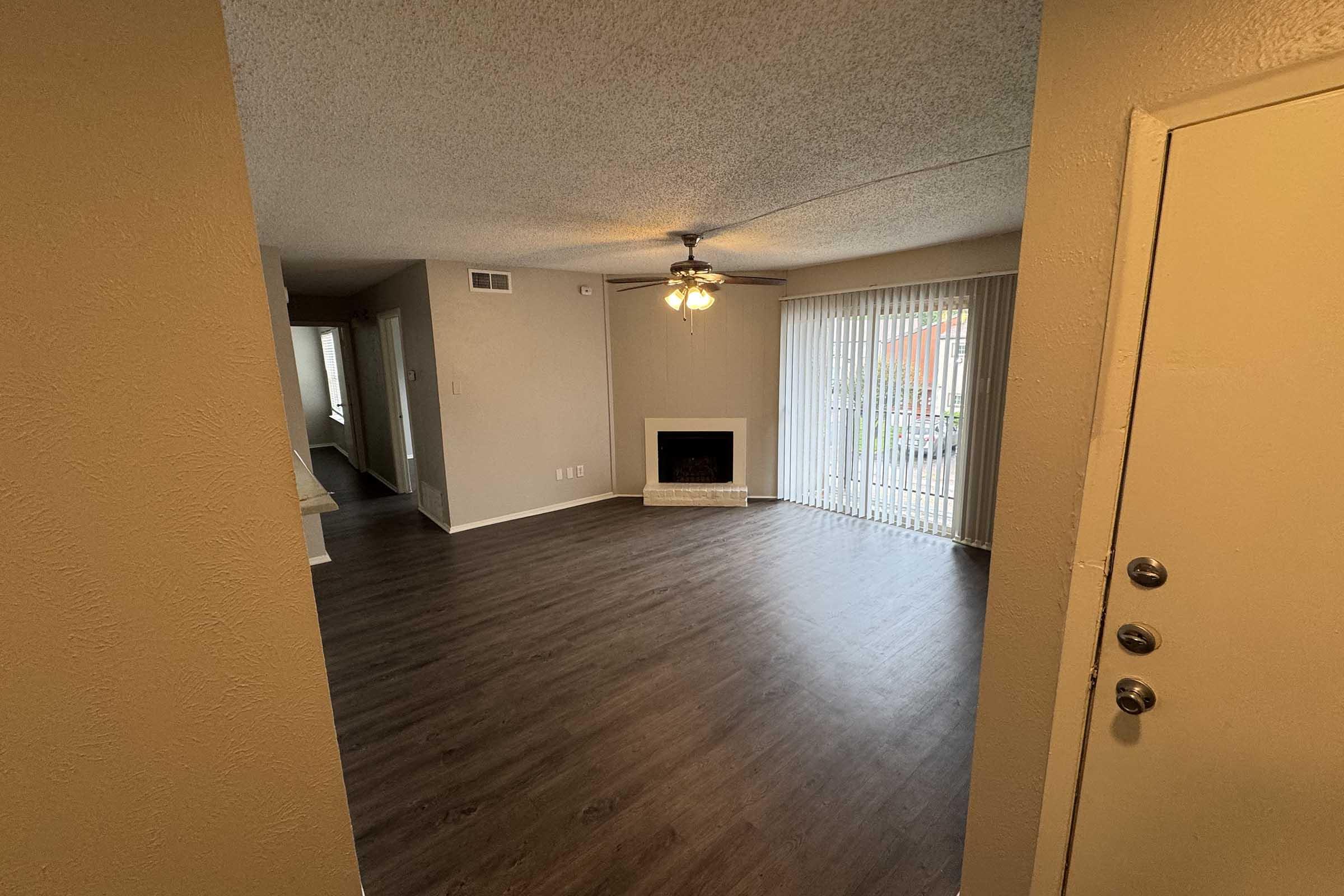 View of a spacious living room featuring a ceiling fan, a fireplace set in a beige wall, and large sliding glass doors with vertical blinds. The floor is covered in dark wooden laminate, and there are light-colored walls and a doorway leading to another room.