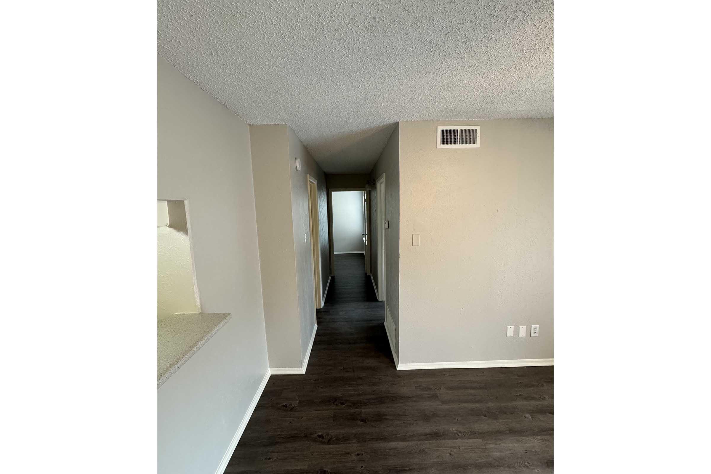 A view of a hallway inside an apartment, featuring light gray walls and a textured ceiling. The flooring is dark wood. On the left, a partially visible kitchen area opens into the hallway. Two doorways are visible, leading to other rooms, with natural light coming from the end of the hallway.