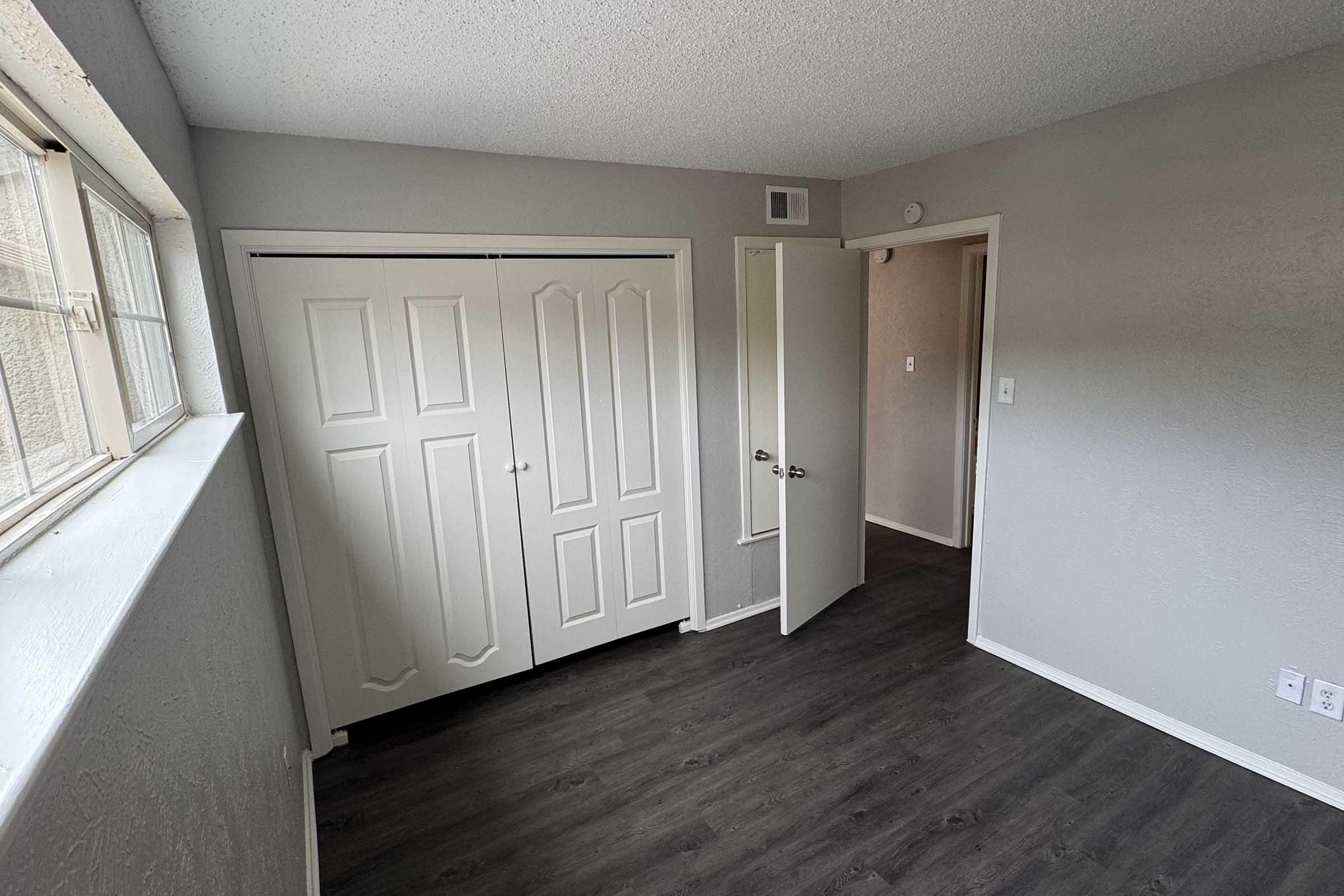 A vacant room featuring beige walls, a large window letting in natural light, and dark laminate flooring. There are double white closets with sliding doors and a partially open door leading to another room. The space is well-lit and simple, ideal for various uses.