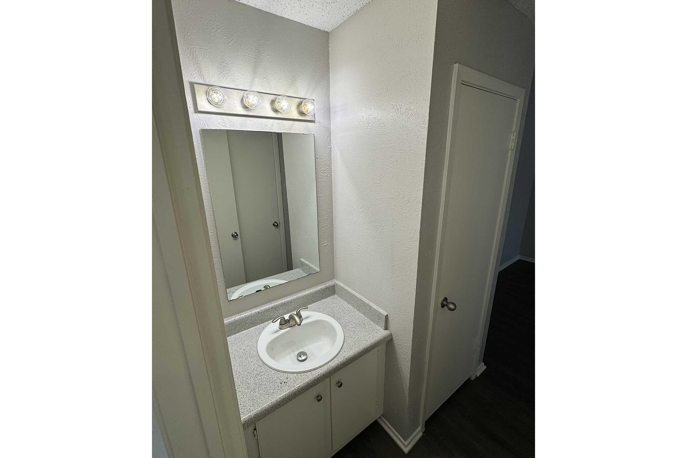A small bathroom area featuring a countertop with a sink, a mirror above, and a light fixture. The walls are painted in a light color, and there is a door on the right leading to another room. The floor is dark, contrasting with the lighter shades of the room.