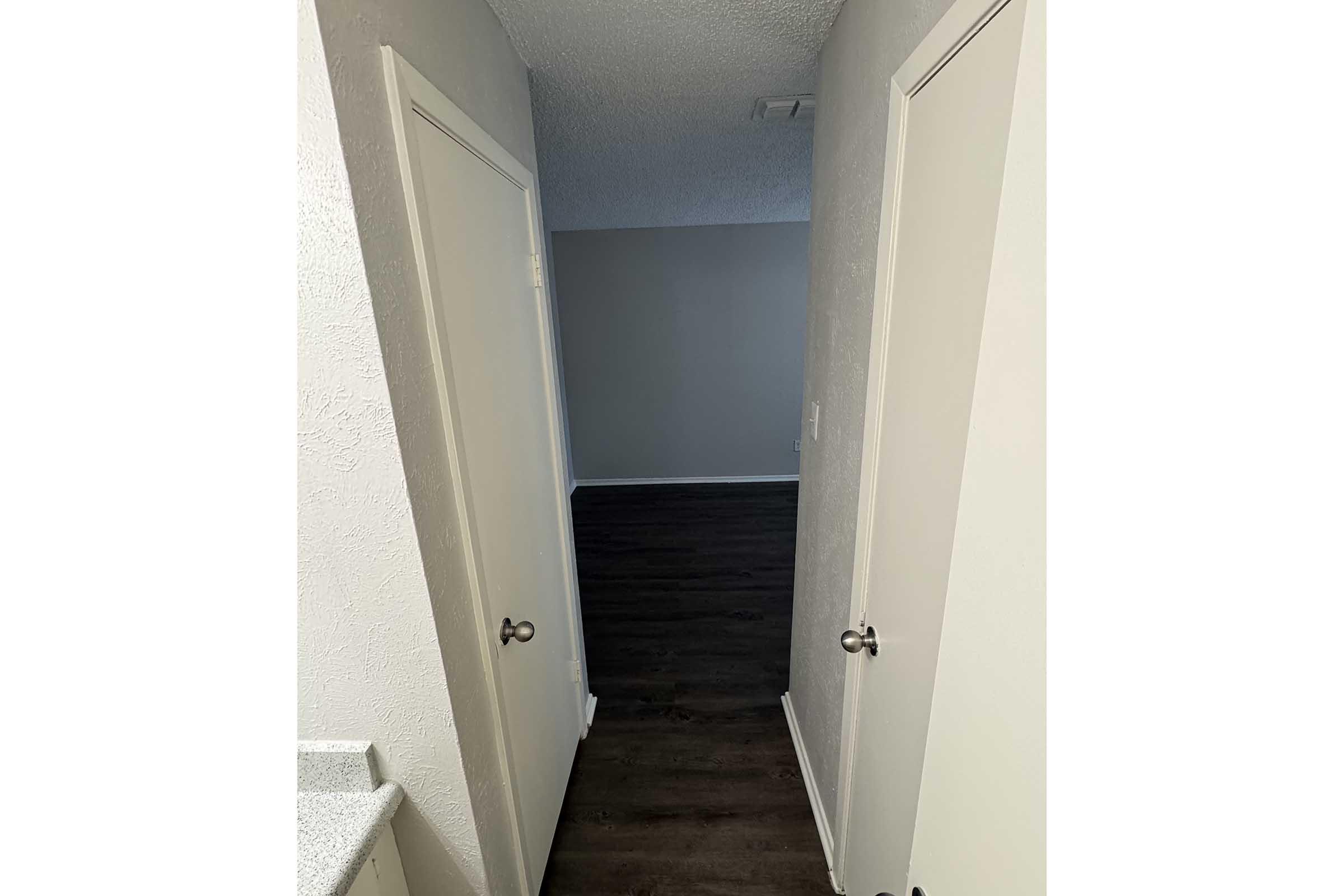 A narrow hallway with two closed doors on the left and a view of a spacious room with wood-like flooring in the background. The walls are painted in a light color, and the ceiling appears textured.