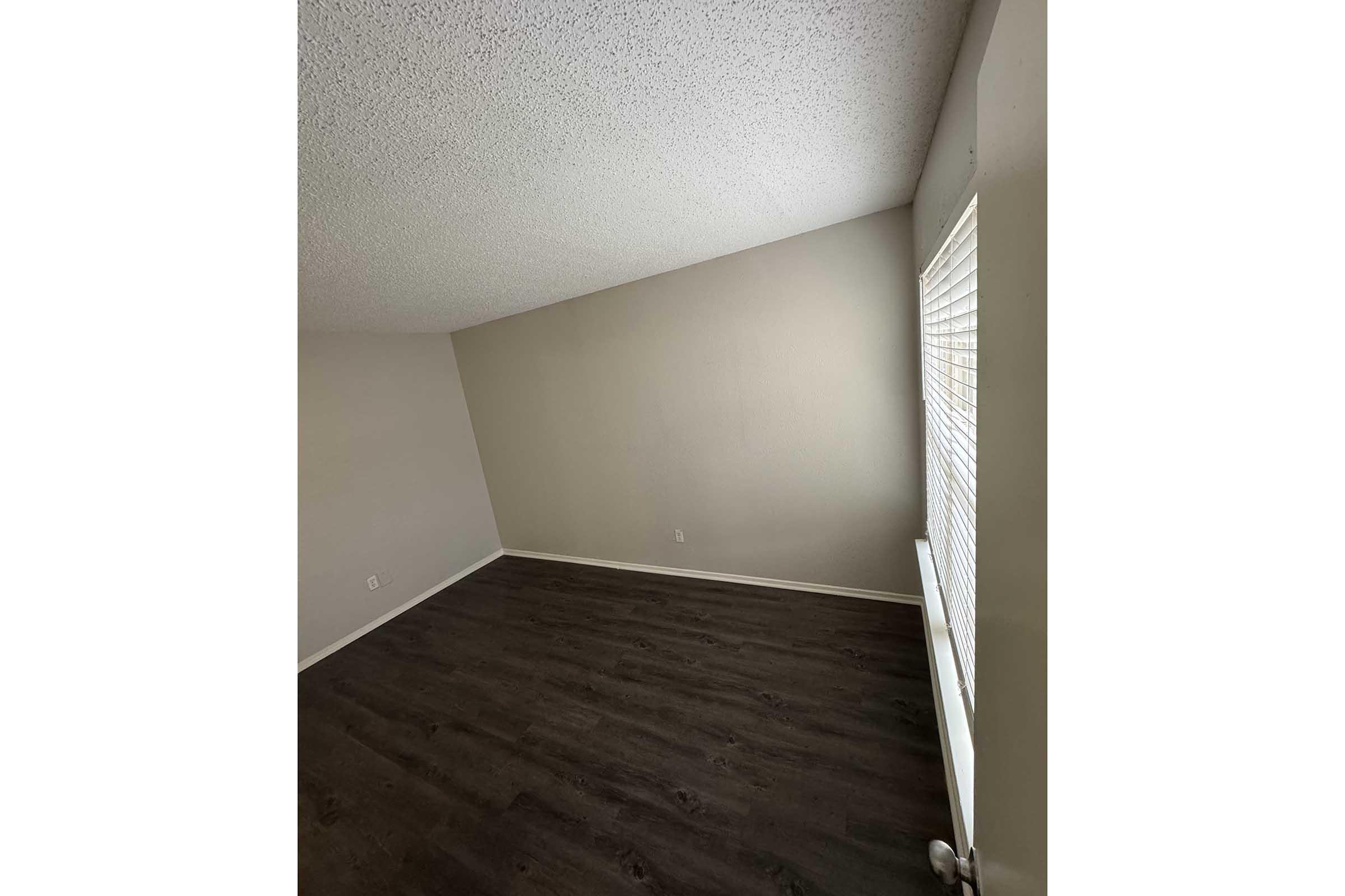 An empty room featuring light gray walls and a textured ceiling. The floor is dark wood, and there is a window with blinds on the right side, allowing natural light to illuminate the space. The room appears clean and uncluttered, suitable for various uses or personal customization.
