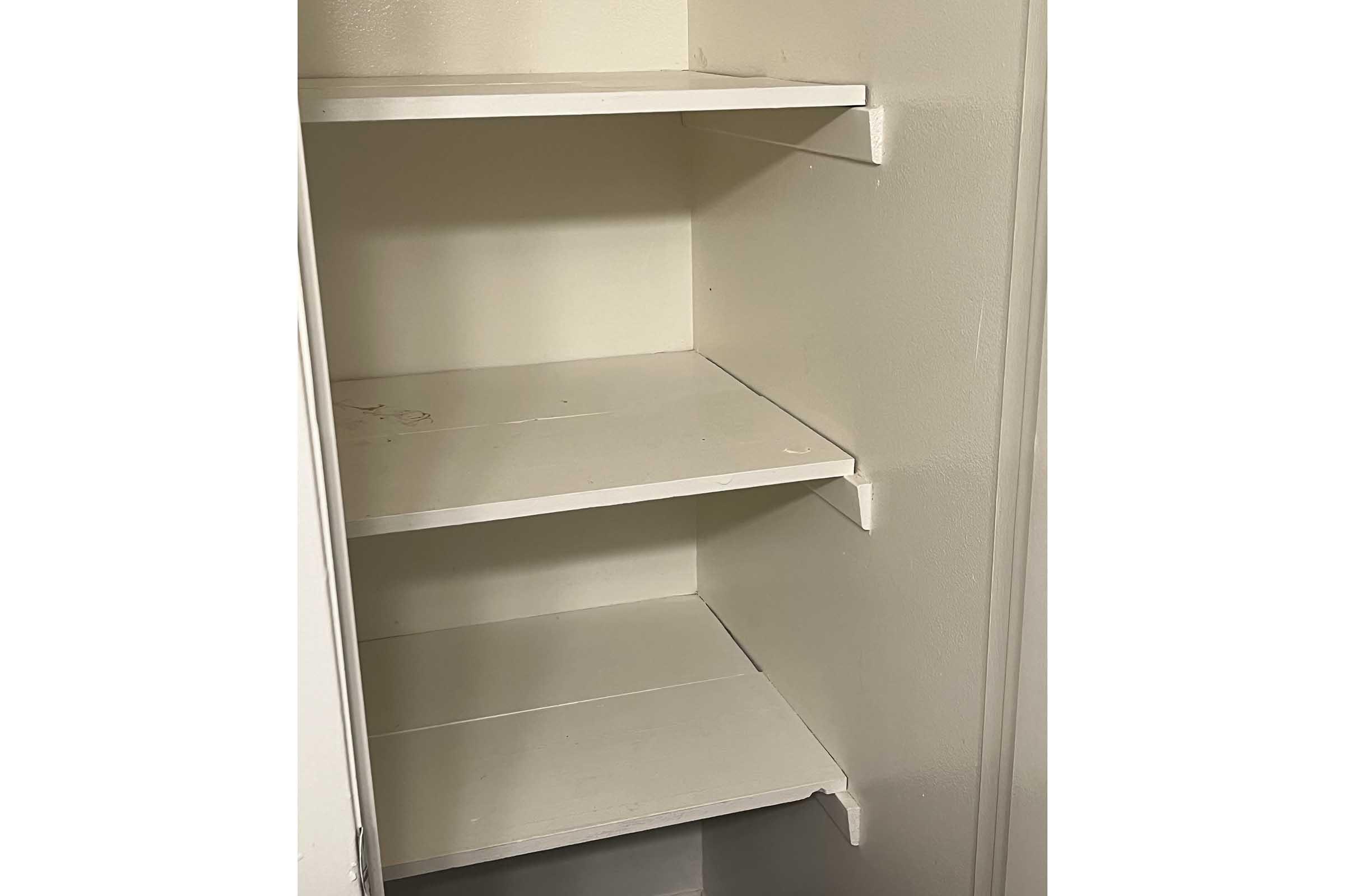 Empty shelving unit with three white, flat shelves against a light-colored wall. The shelves have a smooth surface and are positioned inside a closet or pantry space, with some minor marks visible on the shelves. The background is neutral, creating a simple and uncluttered appearance.