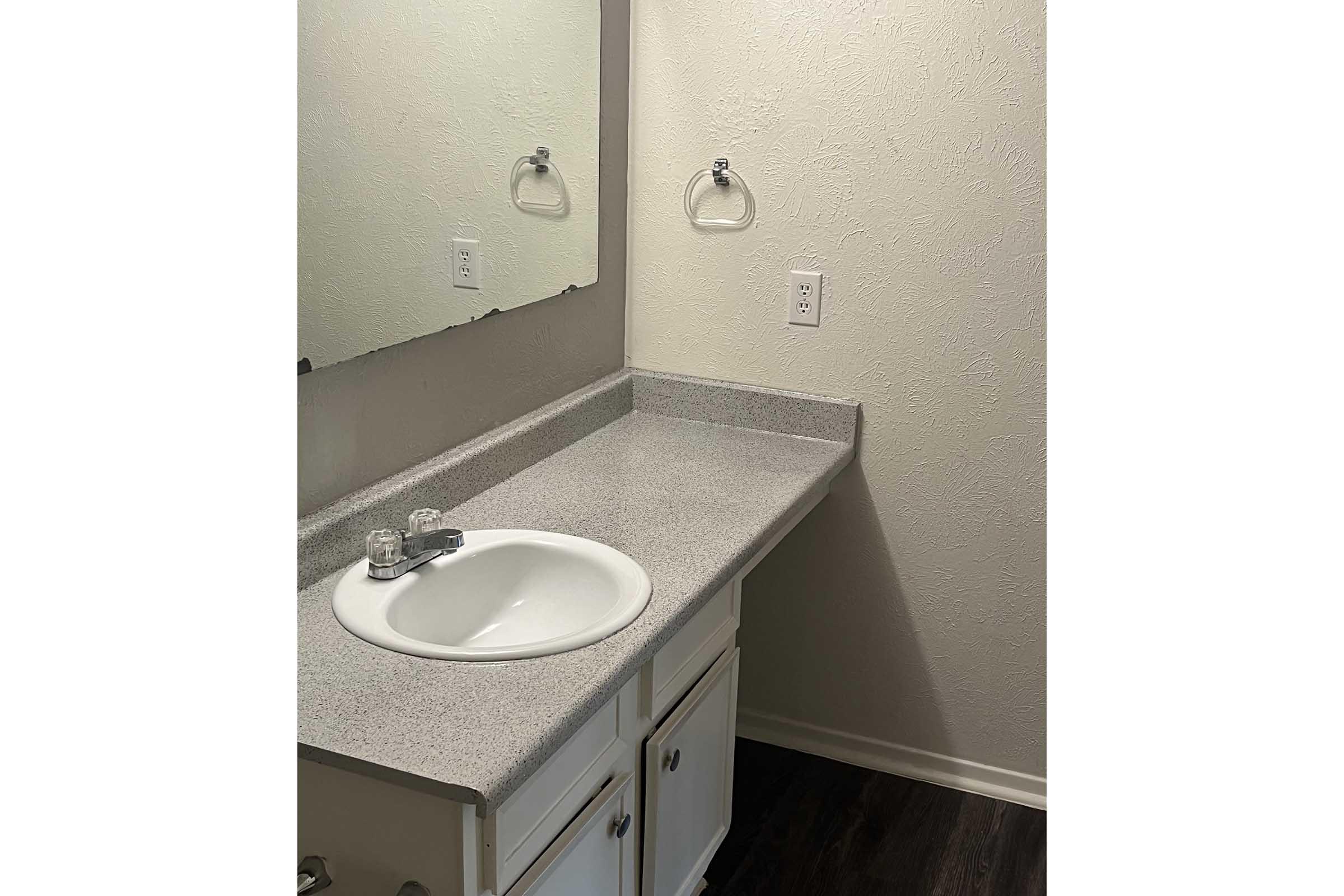 A clean bathroom with a simple countertop, a sink with a faucet, and an empty space for toiletries. The wall features a large mirror and a towel rack. The surrounding walls are painted in a light color, complementing the dark flooring.