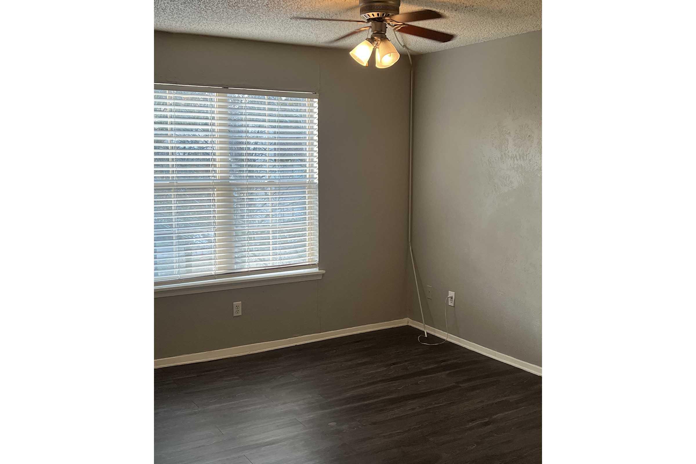 A small, empty room with gray walls and dark wooden flooring. It features a window with white blinds allowing natural light, and a ceiling fan with two bulbs. There is a power outlet on the wall, and the space is minimalistic, indicating it may be ready for decoration or furniture.
