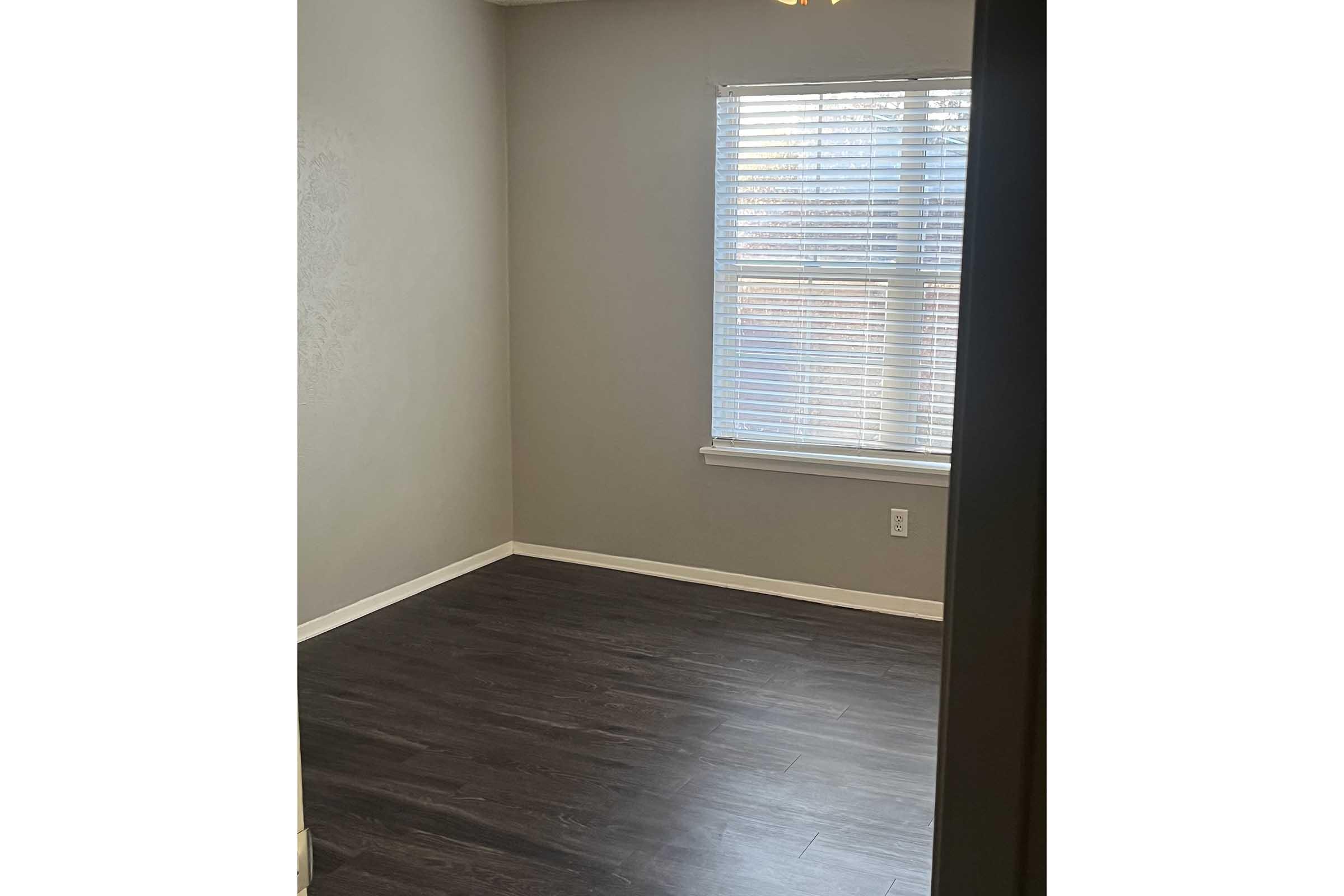 A small, empty room with gray walls and a large window featuring white blinds. The floor is dark laminate wood. There are no furnishings or decorations, creating a minimalist appearance. Natural light enters through the window, illuminating the space.