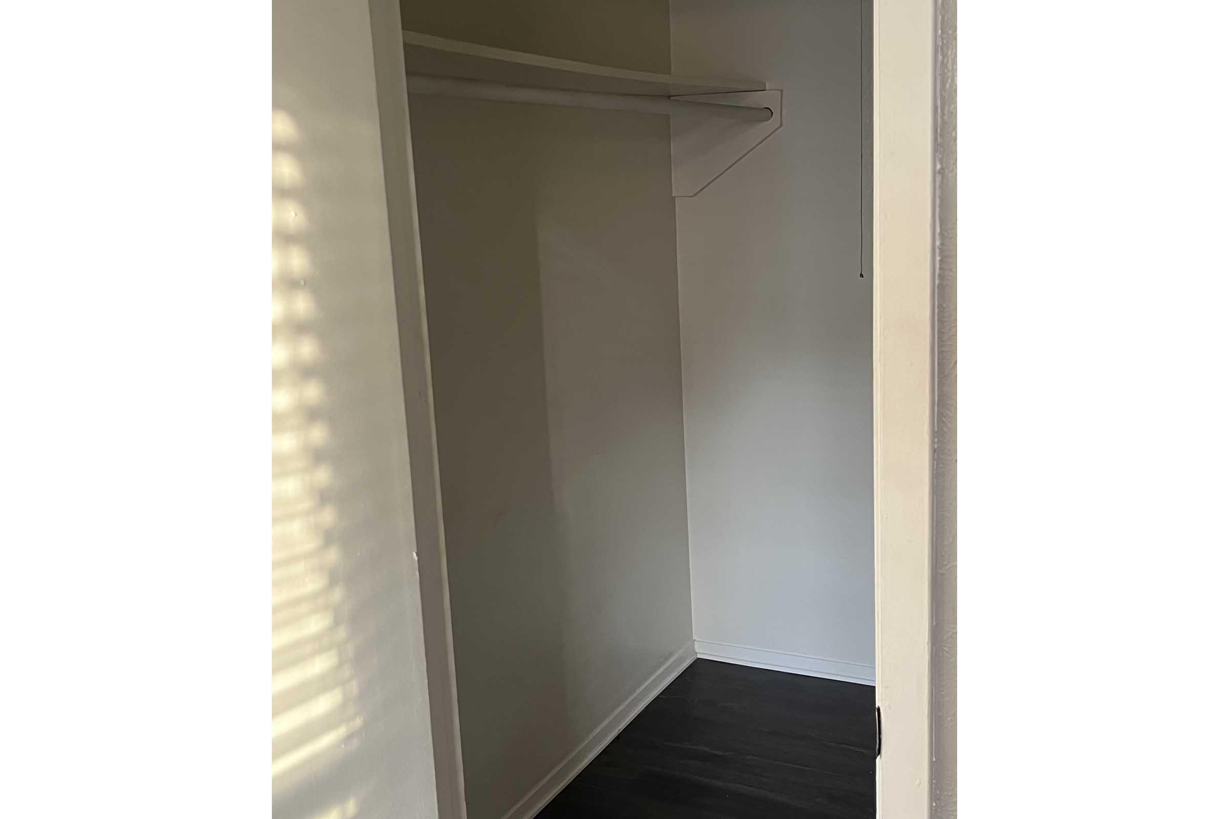 A small, empty closet with a white wall, featuring a single shelf above for storage. The floor is dark, and there is a faint shadow from light coming through a nearby window.