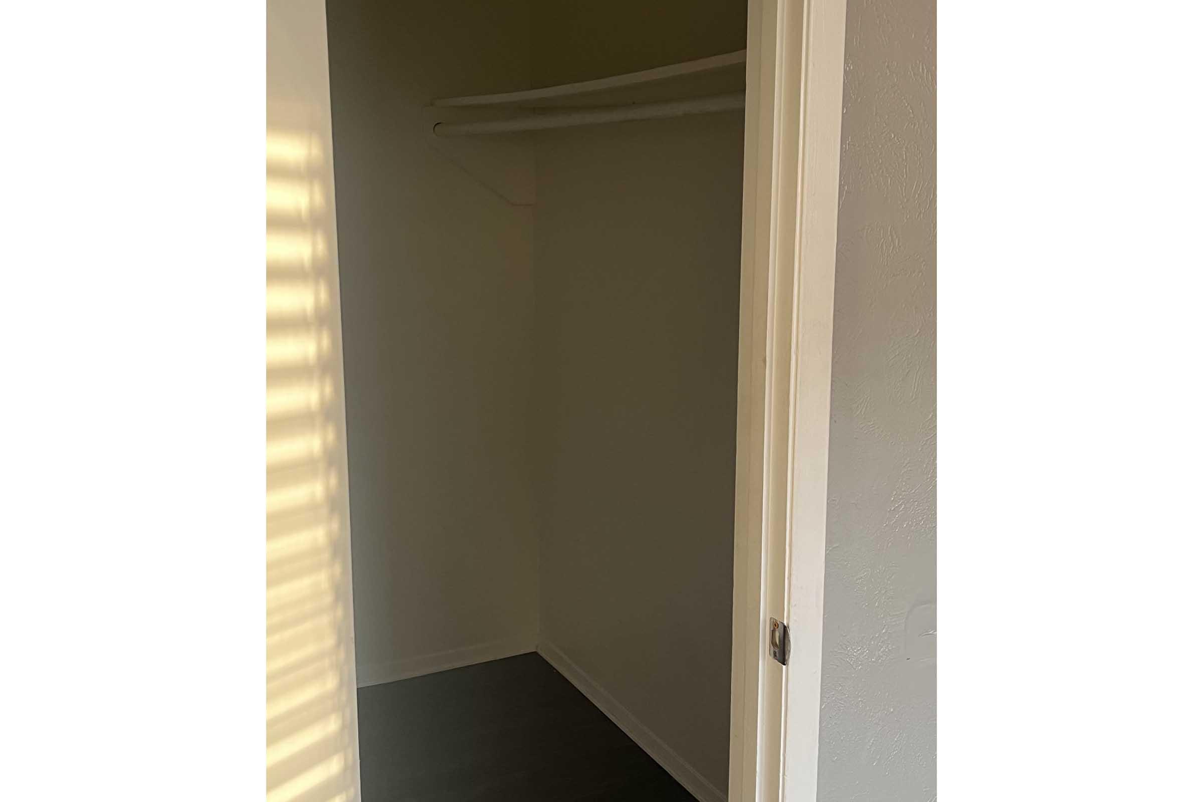 A small, empty closet with a single shelf along the upper wall. The walls are painted light, and sunlight casts shadows through a nearby window. The floor appears dark, contrasting with the light-colored walls. The open door reveals the closet space.