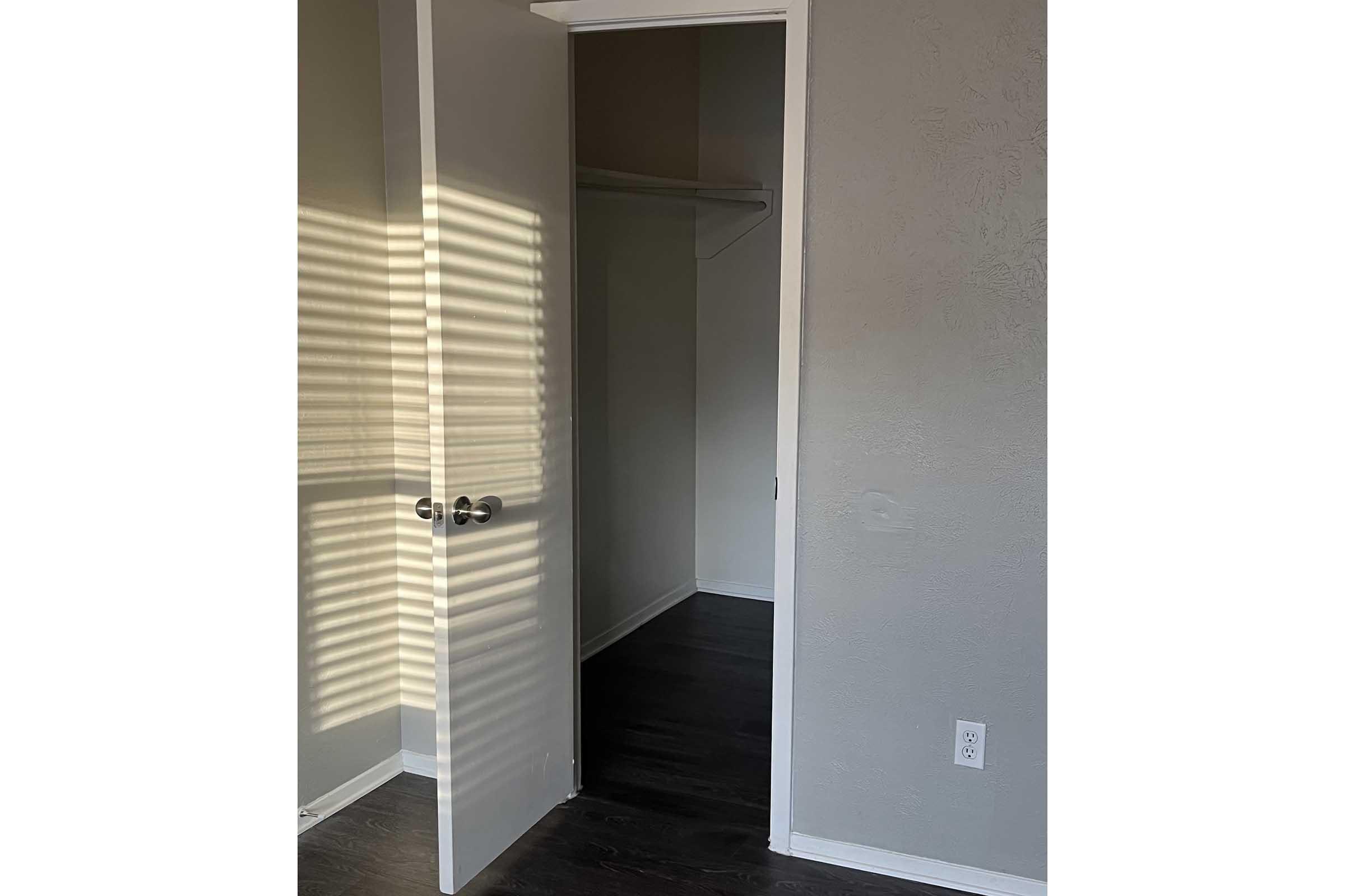 A partially open door leads into a small room with gray walls and a closet on the left side. Soft sunlight casts stripes on the floor from the door frame, while dark wooden flooring is visible inside the room. The overall atmosphere is calm and inviting.