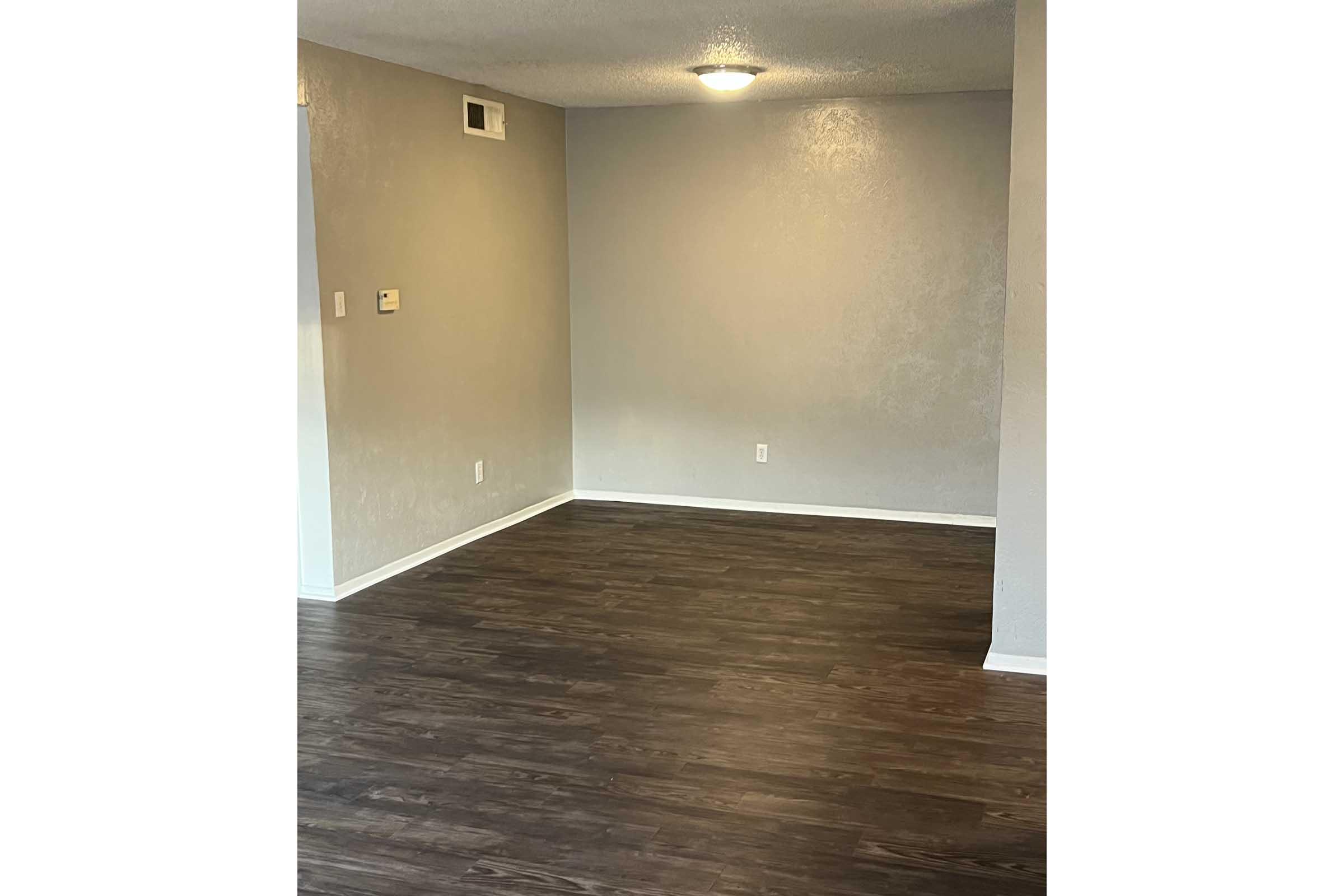A spacious, empty room with light gray walls and dark brown laminate flooring. The ceiling is illuminated by a round light fixture, and there are electrical outlets on the walls. The room appears clean and ready for furnishing.