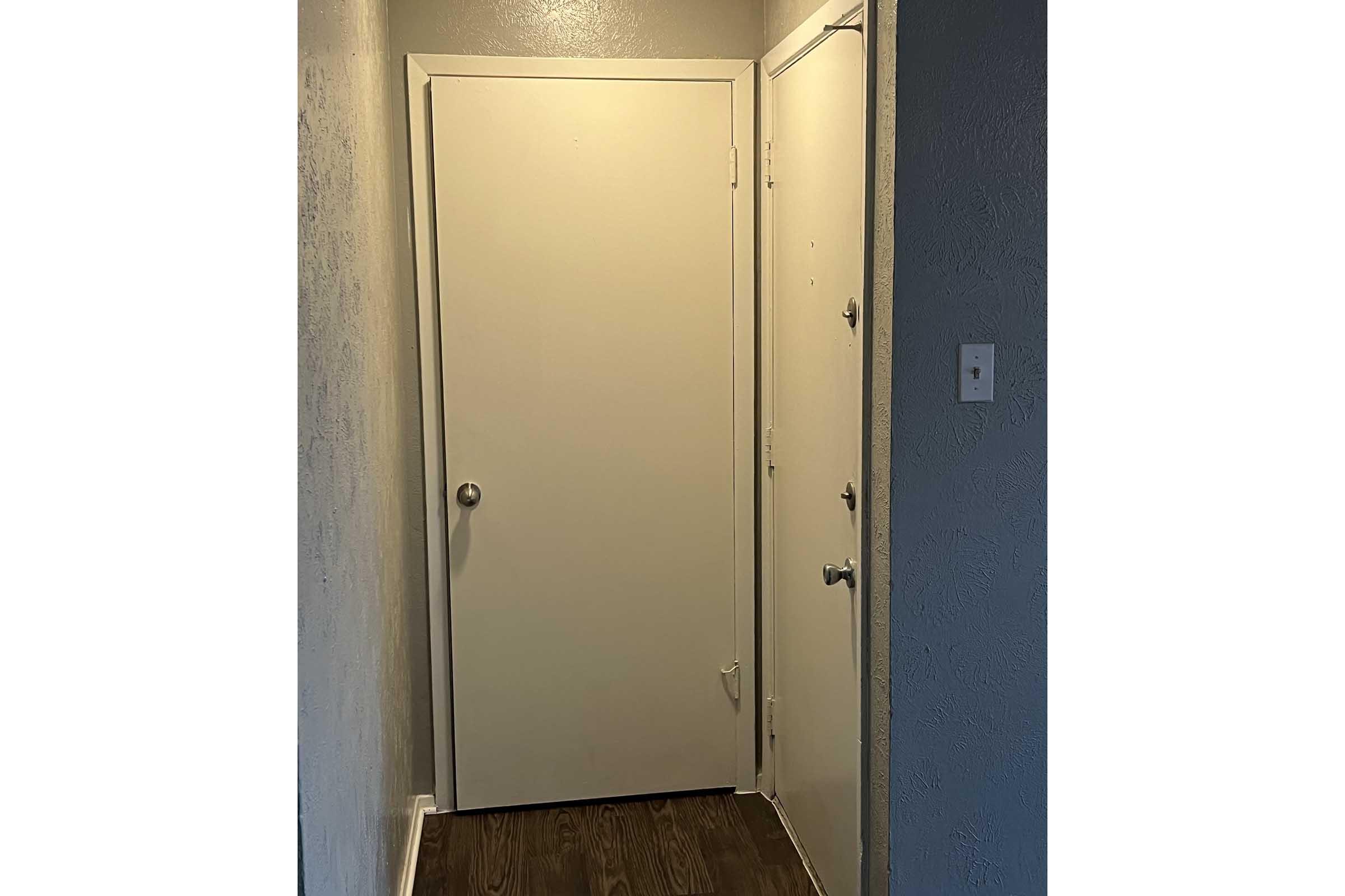 A narrow hallway featuring a light-colored wall and a wooden floor. Two doors are visible: one is a plain white door on the left, and another door with a doorknob on the right. There is also a light switch on the wall near the right side. The overall lighting is bright and neutral.