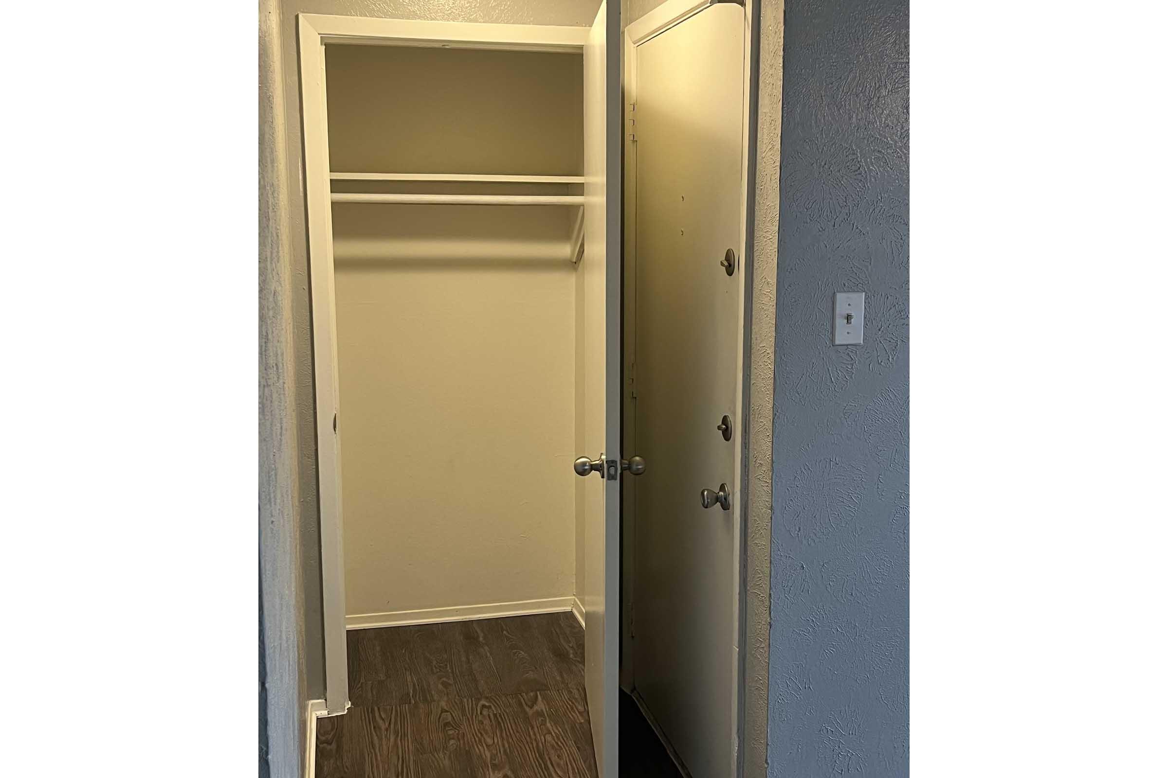 A narrow hallway leading to an empty closet on the left and a door on the right. The closet features a single shelf above a wooden floor. The door is partially visible, with multiple doorknobs and no visible details. The walls have a textured finish in a light color.