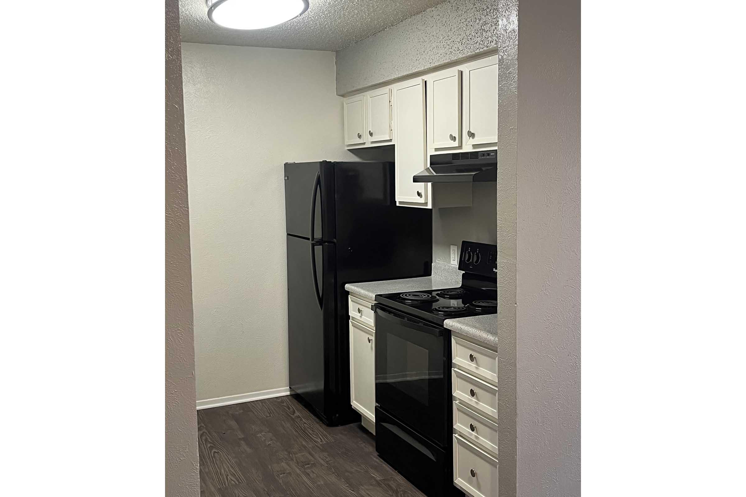 A small kitchen featuring white cabinets and a black refrigerator; there's a black stove and oven, along with a gray countertop. The flooring is wood-like, and a ceiling light illuminates the space. The kitchen appears modern and compact, suitable for cooking and food preparation.