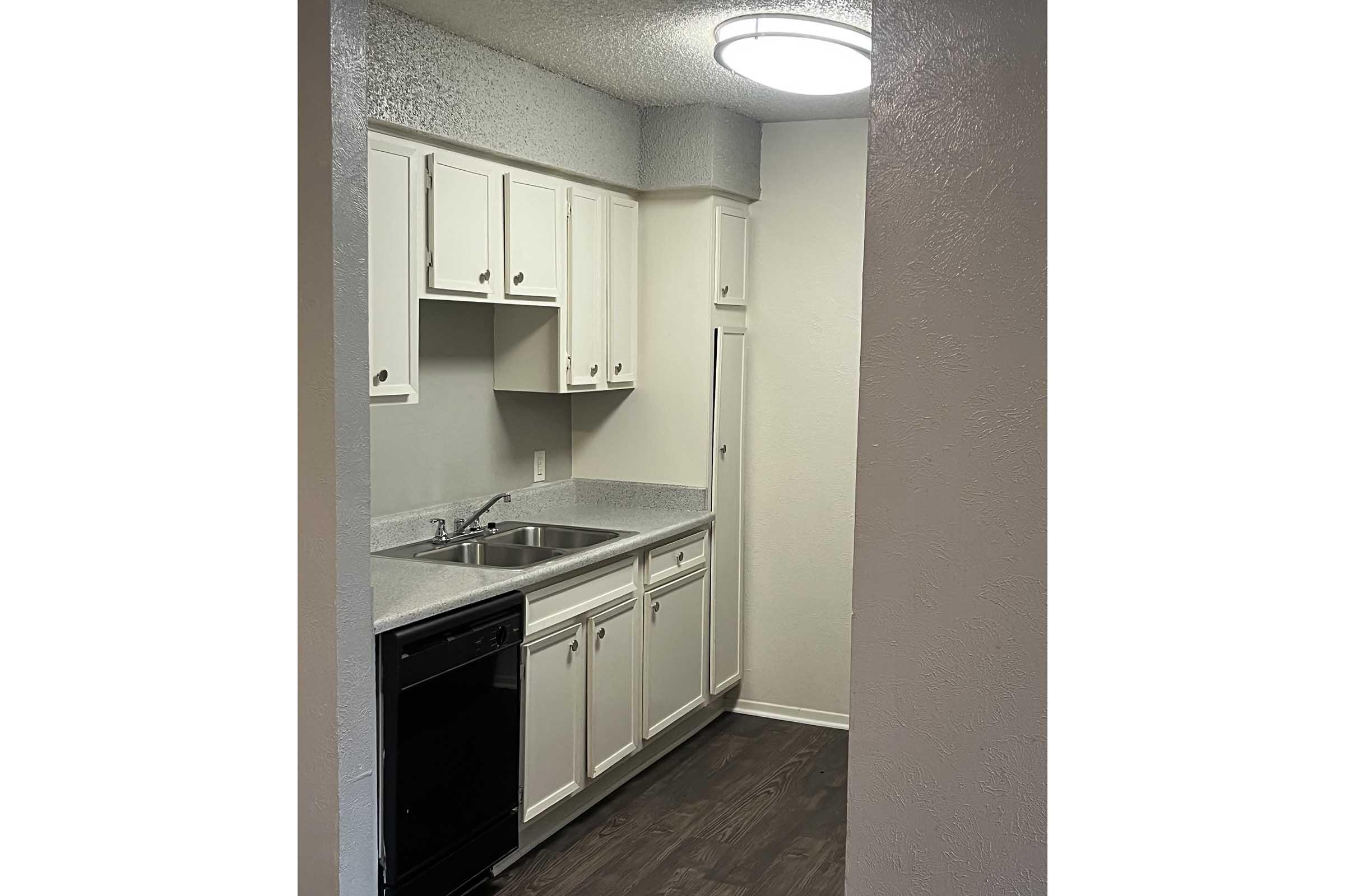 A small kitchen featuring white cabinets, a stainless steel sink, a black dishwasher, and a light-colored countertop. The space includes a wall-mounted light fixture and a portion of a refrigerator visible. The floor is dark wood, providing a modern contrast to the lighter cabinets.