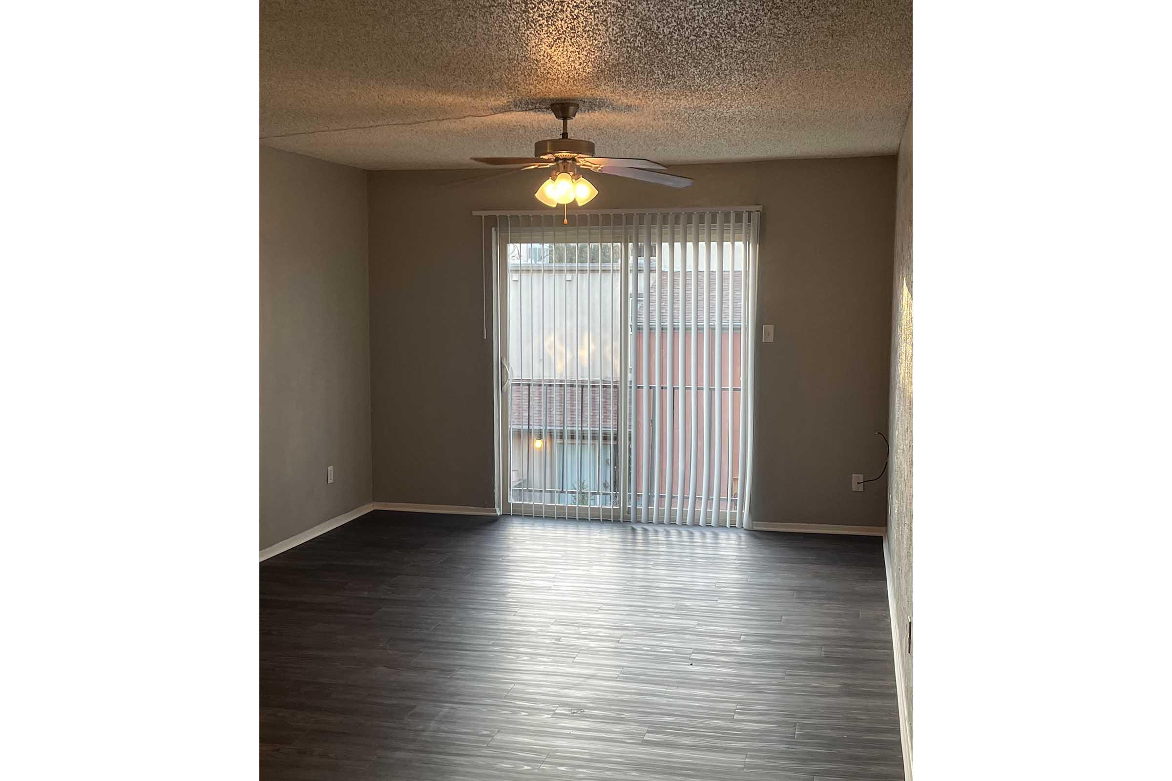 A well-lit room with a textured ceiling, featuring a ceiling fan and a sliding glass door with vertical blinds. The floor is made of dark, polished laminate. Light gray walls complement the natural light coming from outside.