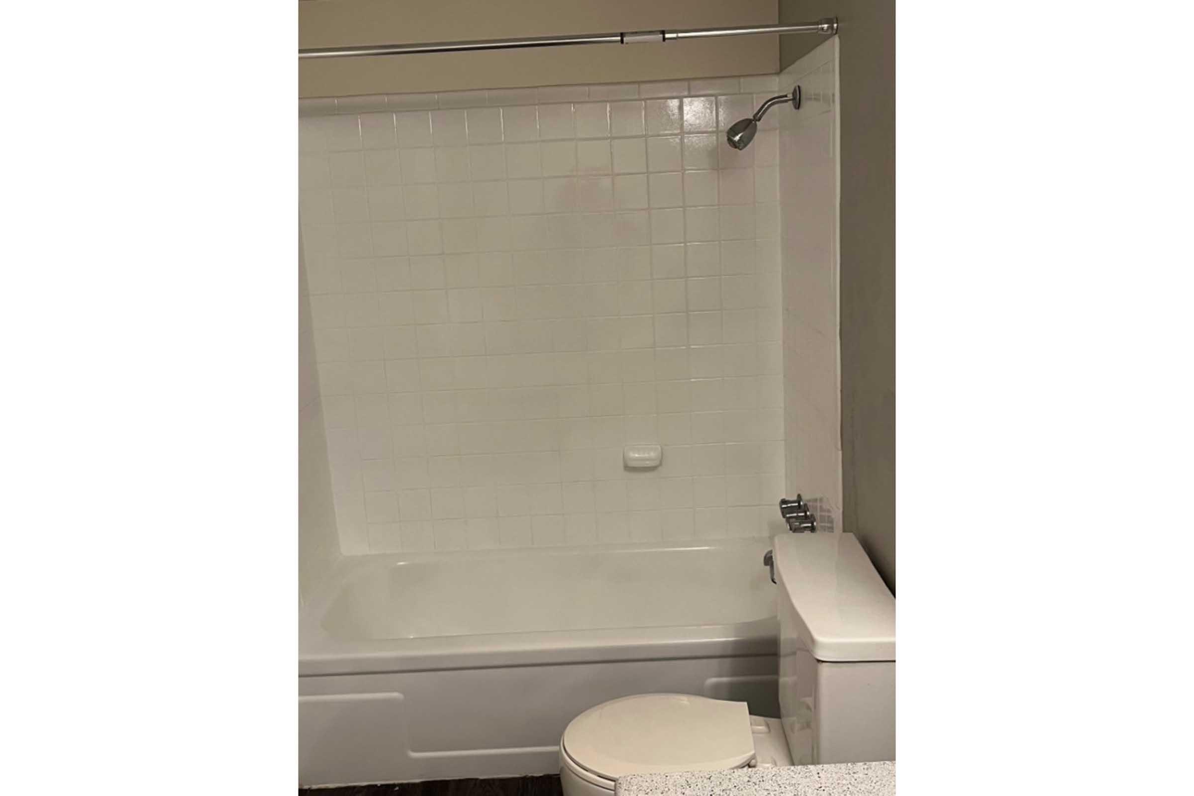 A clean bathroom featuring a white tub with a showerhead above, surrounded by white tiled walls. There's a toilet adjacent to the tub and a countertop with a light-colored surface, giving the space a simple and modern aesthetic.