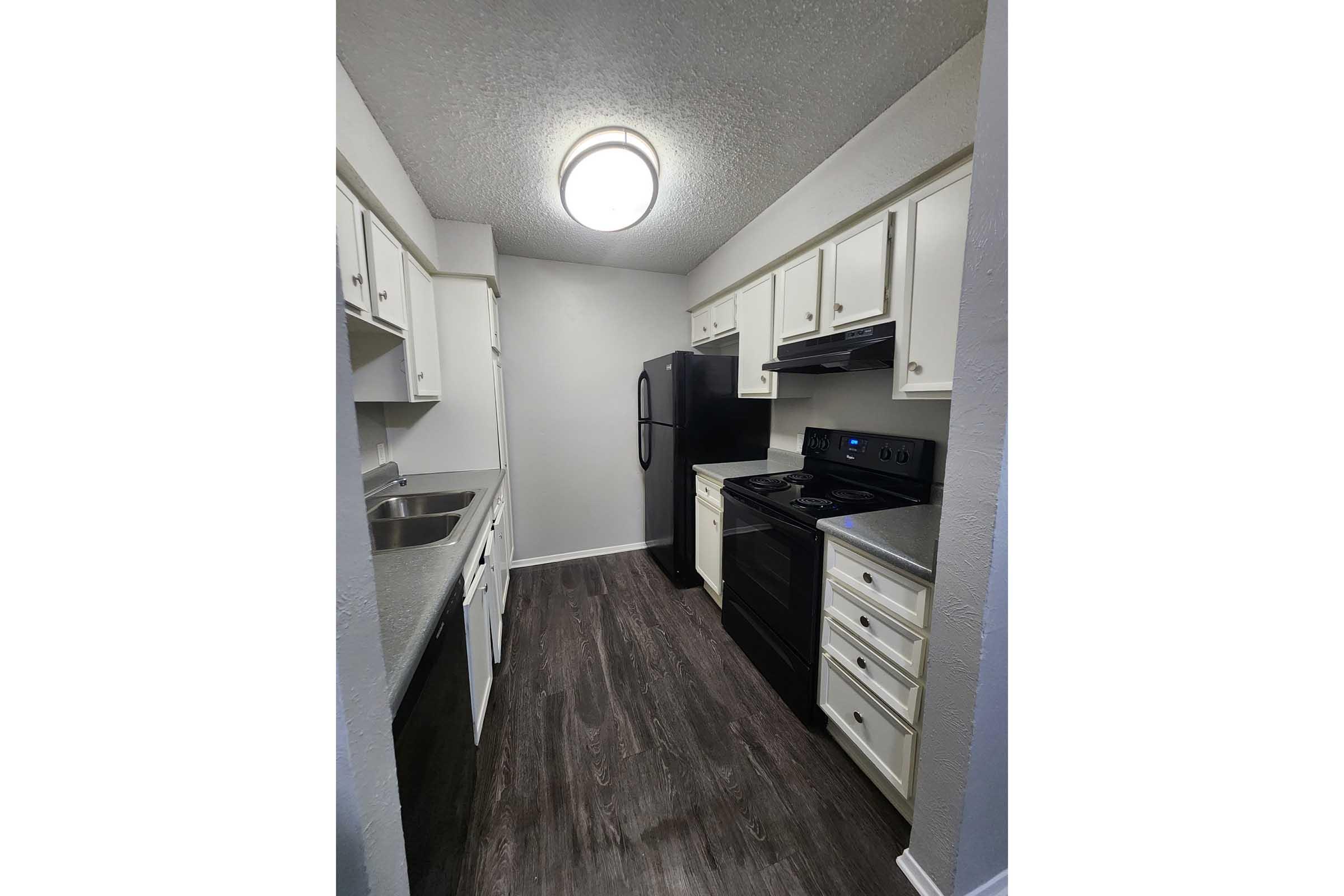 A modern kitchen with white cabinets, black appliances including a refrigerator and stove, a stainless steel sink, and a light fixture. The floor is dark wood laminate, creating a contemporary look. The space is well-lit and organized.