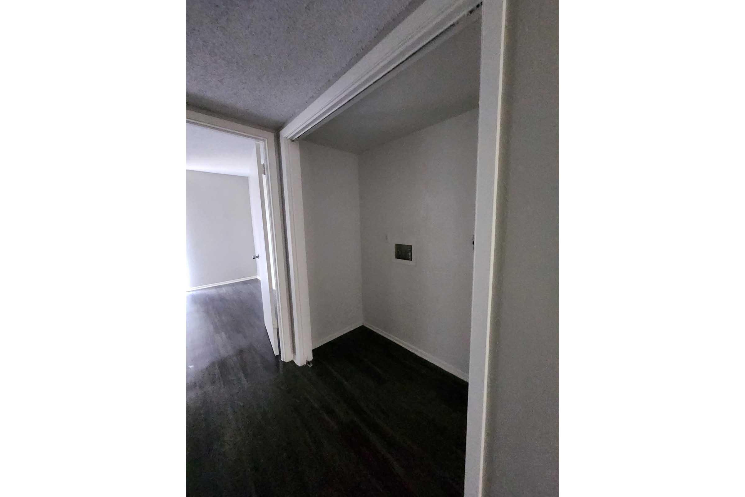 A narrow hallway leads to a dimly lit room with white walls. There is a doorframe on the left side, and a blank wall with an electrical outlet is visible. The flooring appears dark, adding contrast to the light-colored walls. The overall atmosphere is quiet and minimalistic.