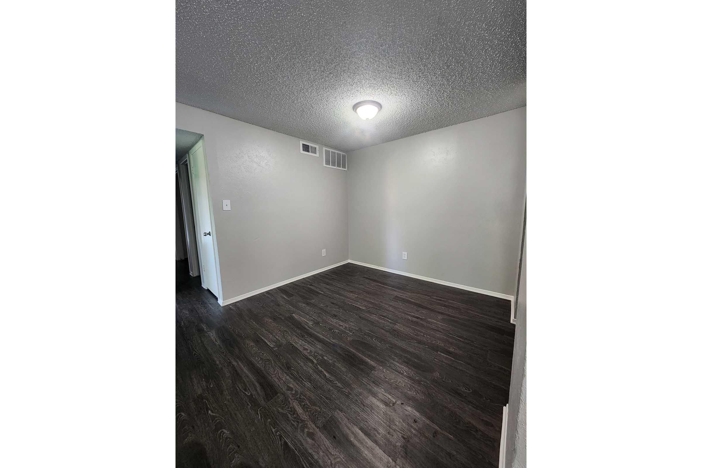 Empty room with gray walls and a textured ceiling. The floor is dark wood, and there is a ceiling light fixture. A door is visible on the left side, leading to another space. No furniture or decorations are present, giving the room a clean, minimalist appearance.
