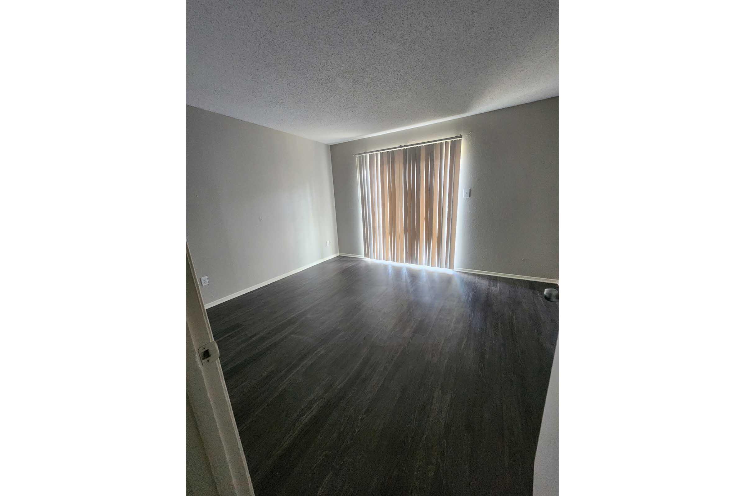 Empty room with dark wood flooring and a large window covered by beige vertical blinds. Soft natural light is coming through the blinds, creating a bright but sparsely furnished space with neutral-colored walls. No furniture or decorations are visible.