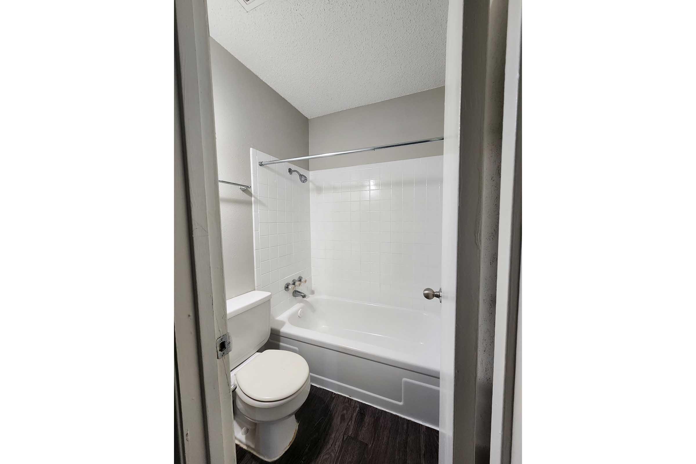 A small bathroom featuring a bathtub with shower head, a toilet, and a sink. The walls are painted in a light color, and there is a sliding glass shower door. The floor appears to be dark-colored laminate. The overall space is well-lit and organized.