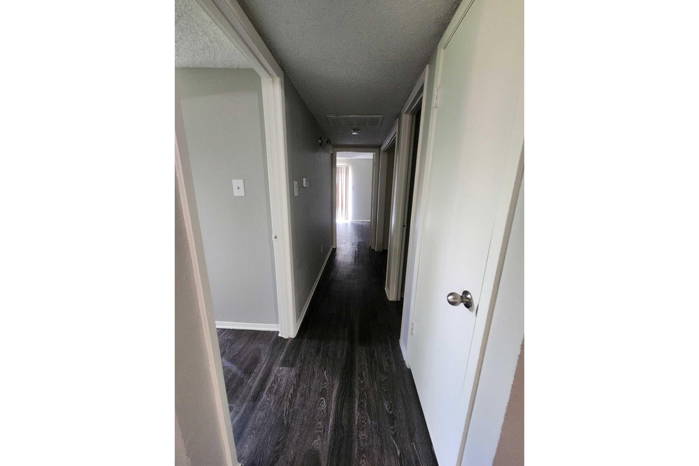 A narrow hallway with light gray walls and dark wood-style flooring. Doors line both sides of the hallway, leading to rooms, while natural light illuminates the far end from what appears to be a window. The space is tidy and modern with a neutral color palette.