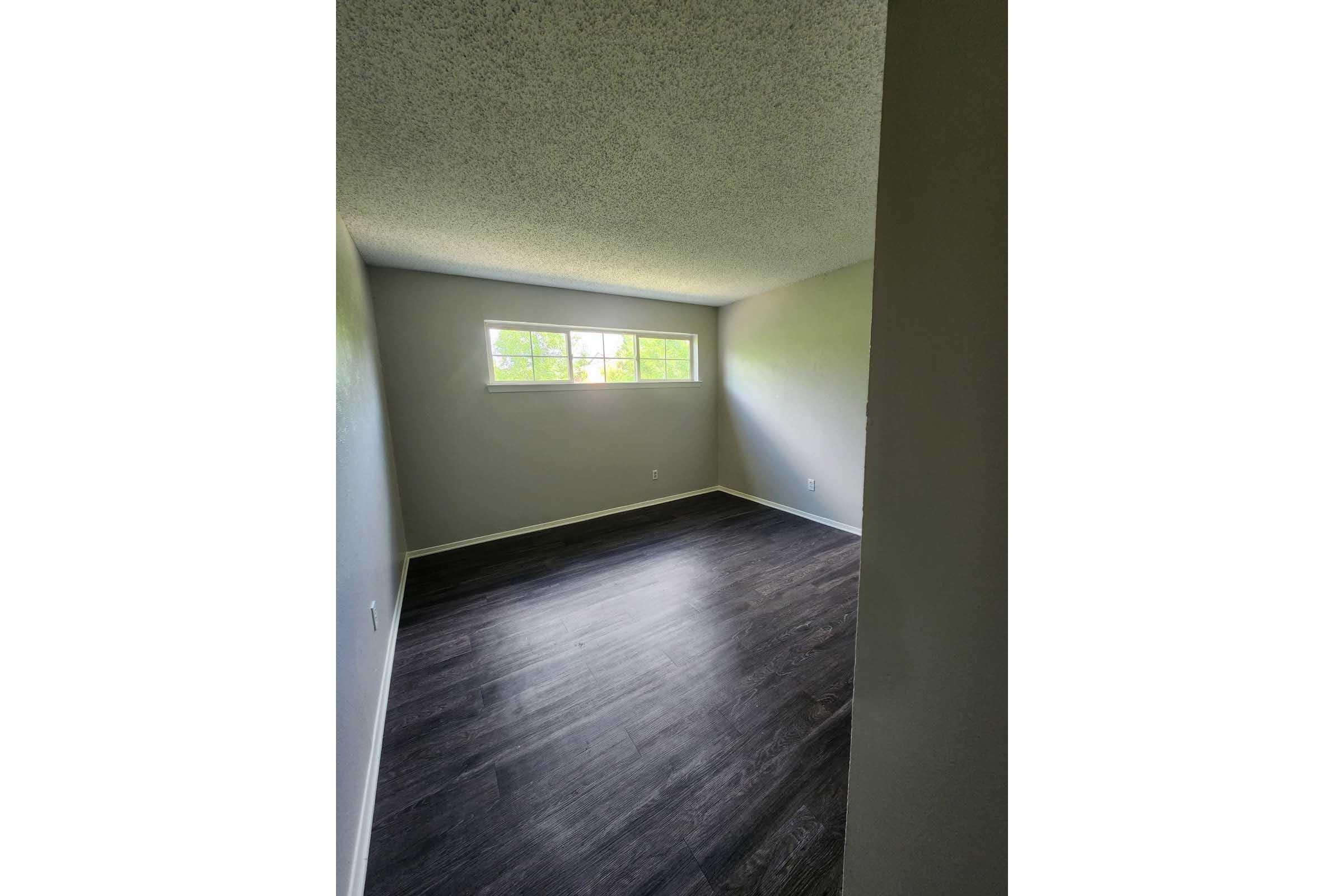 Empty room with gray walls and dark wooden flooring, featuring two windows that allow natural light. The ceiling has a textured finish, and there are no furnishings present, creating a spacious and minimalist appearance.