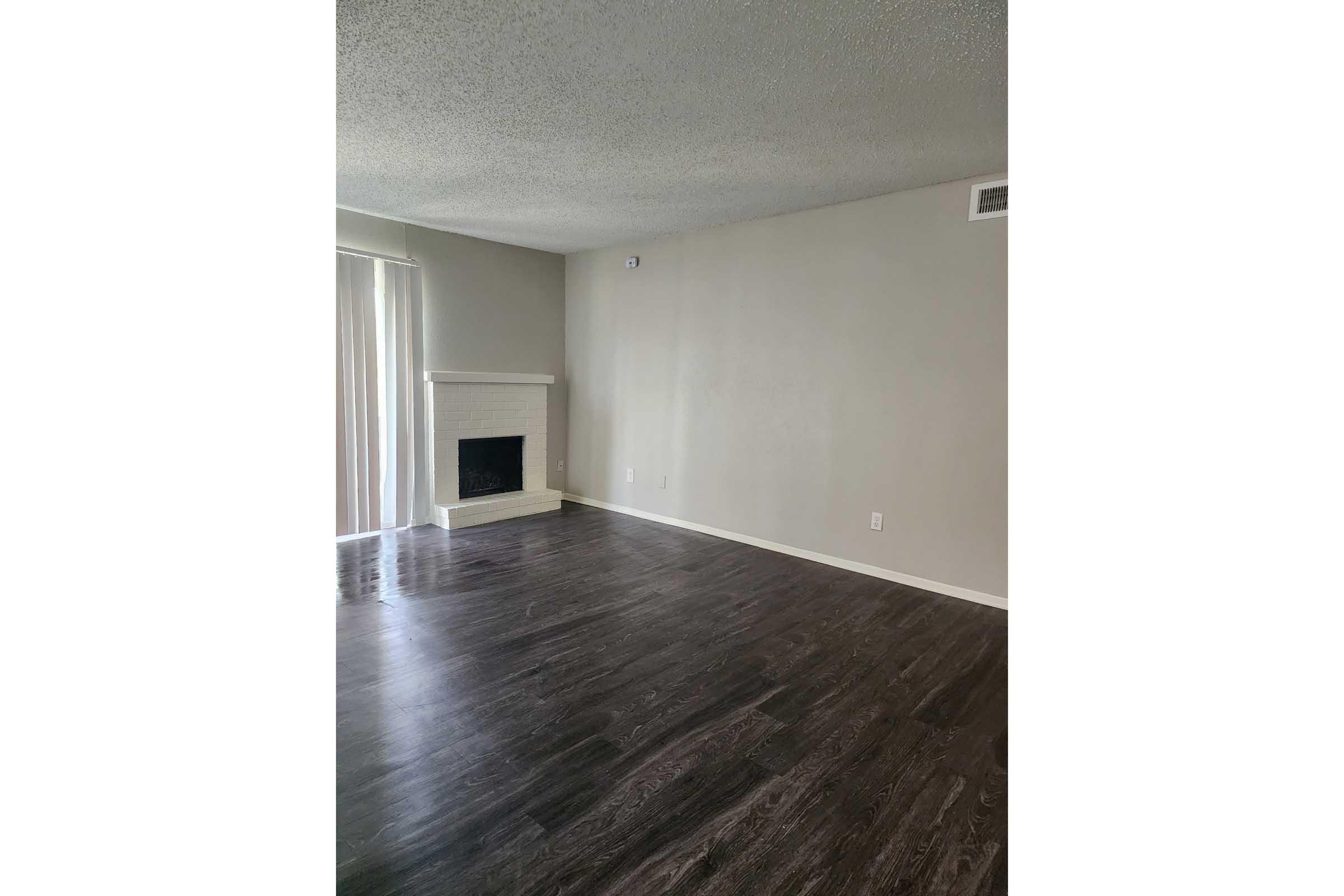 A spacious, empty living room featuring dark hardwood floors and light grey walls. There is a white fireplace on the left side, with a window and sheer curtains allowing natural light to enter. The room is unfurnished and open, creating a minimalist atmosphere.