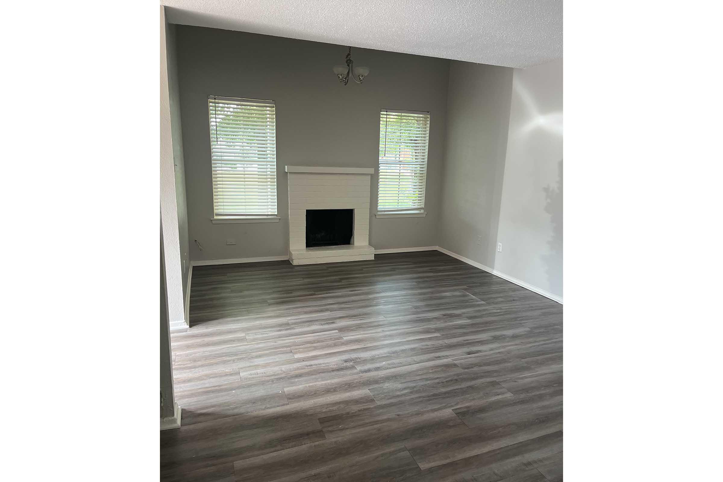 A spacious living room with light-colored walls and a gray accent wall. Two windows with blinds flank a white fireplace. The floor features wood-like laminate in a medium tone, and the room is well-lit by natural light.