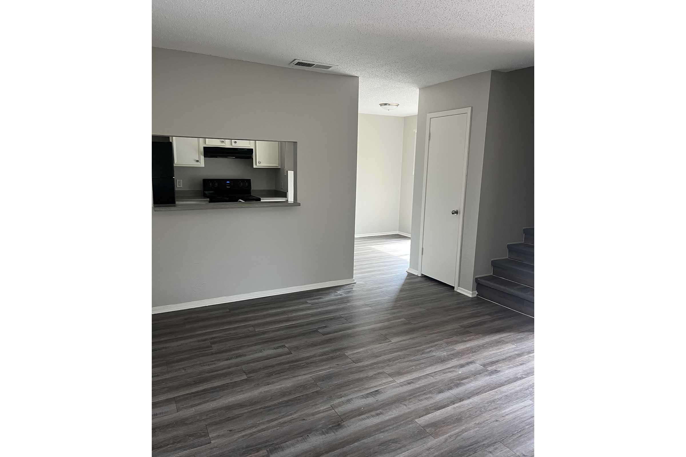 A spacious, modern living area featuring light gray walls and dark laminate flooring. There is a partial view of a kitchen with black appliances and an open design that connects to another room. Stairs lead to a higher level. Natural light appears to enter from an adjacent room.