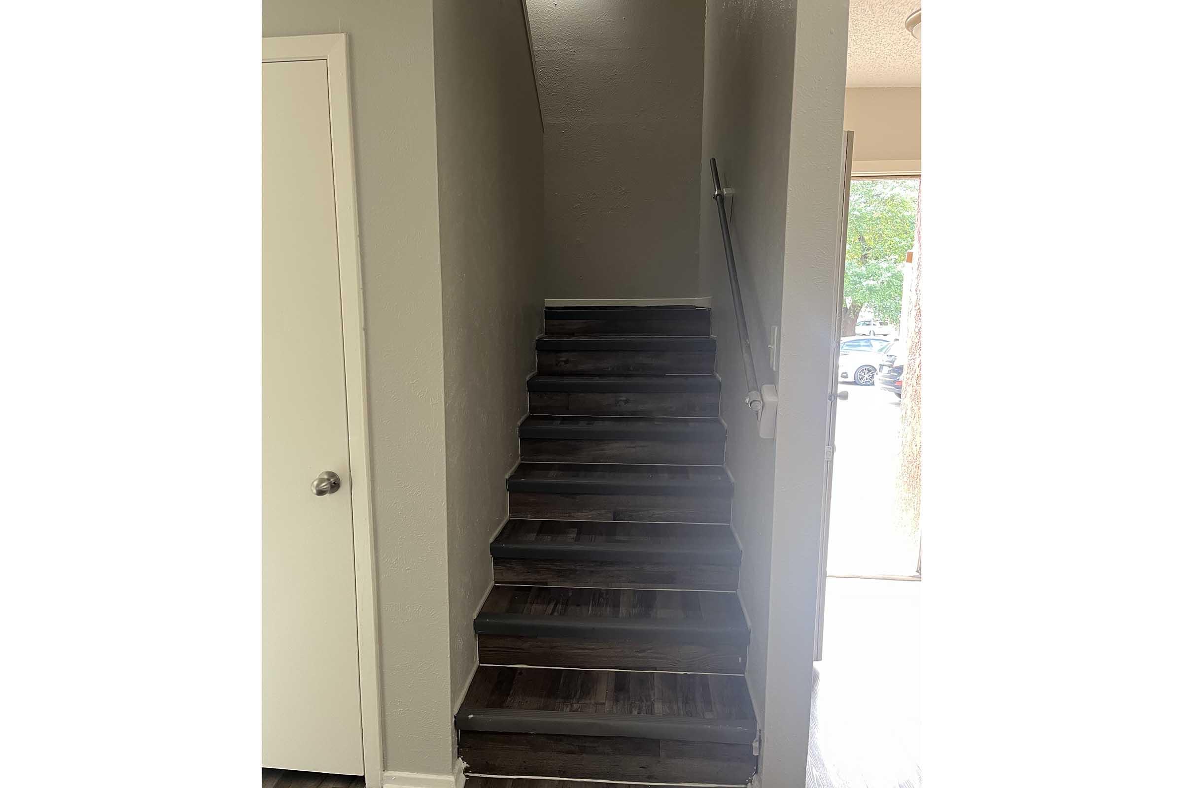 A view of a staircase leading upward, with wooden steps and a light gray wall. There is a door on the left side and a glimpse of natural light from an entrance door at the bottom of the stairs. The overall atmosphere is bright and inviting.