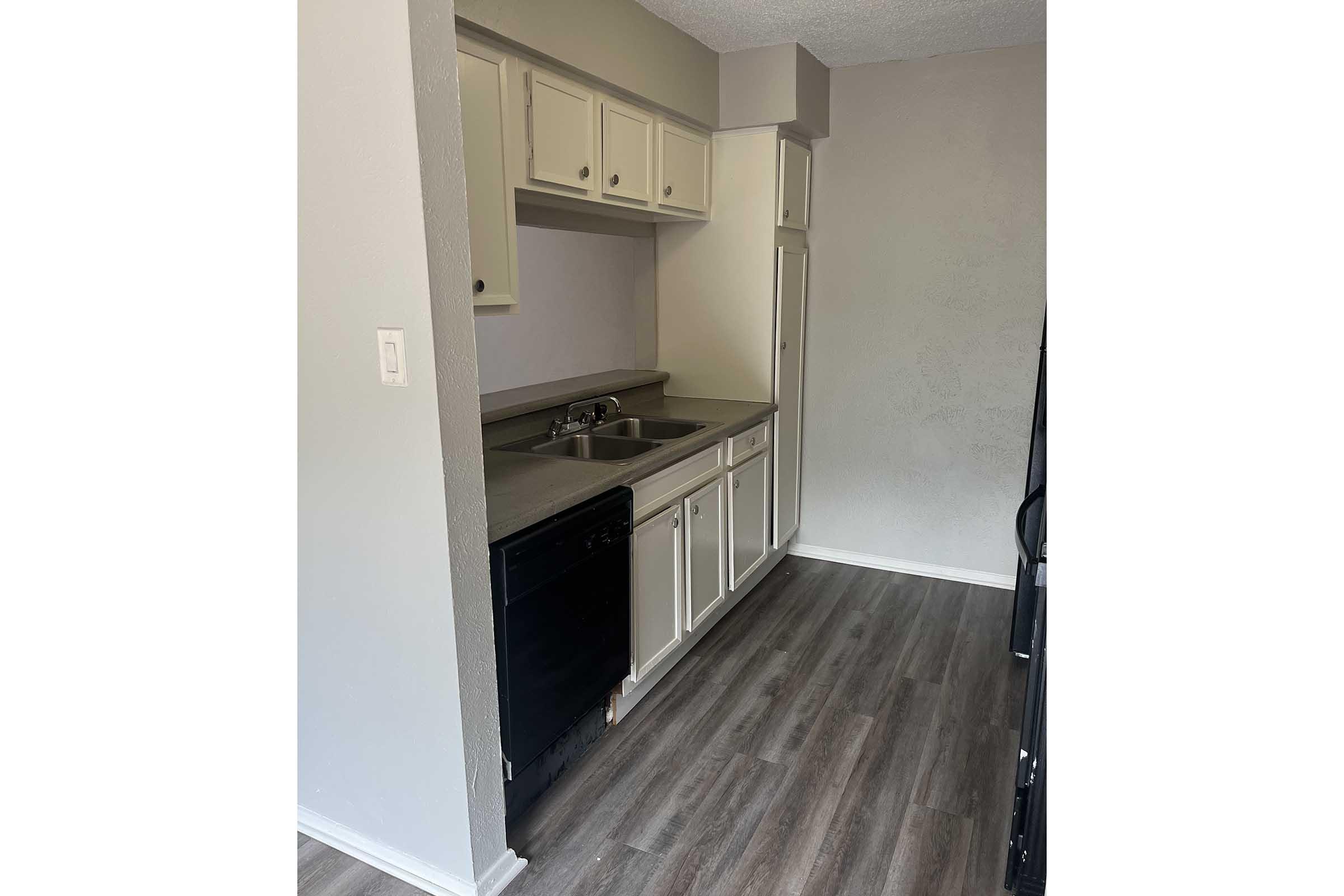A small kitchen featuring light gray walls, beige cabinets, a double sink, and a black dishwasher. The flooring is a wood-like laminate. There is a refrigerator visible in the corner, and the layout suggests ample counter space for cooking and food preparation.