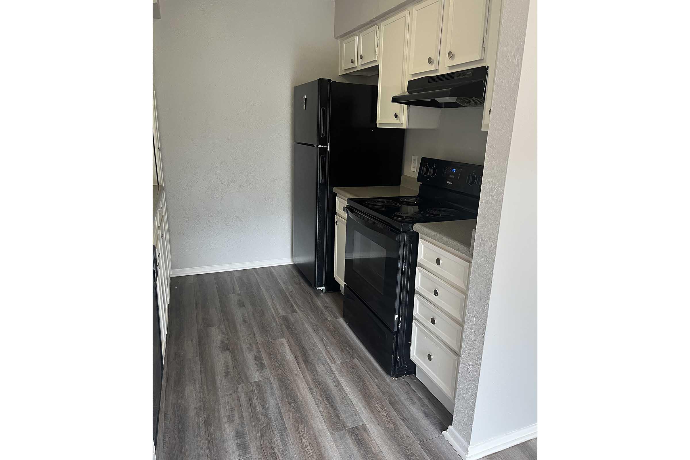A small kitchen featuring white cabinetry, a black refrigerator, black stove and oven, and a matching black microwave. The floor has light-colored wood laminate, and the walls are painted light gray, creating a modern and clean appearance. Natural light comes from the left side.