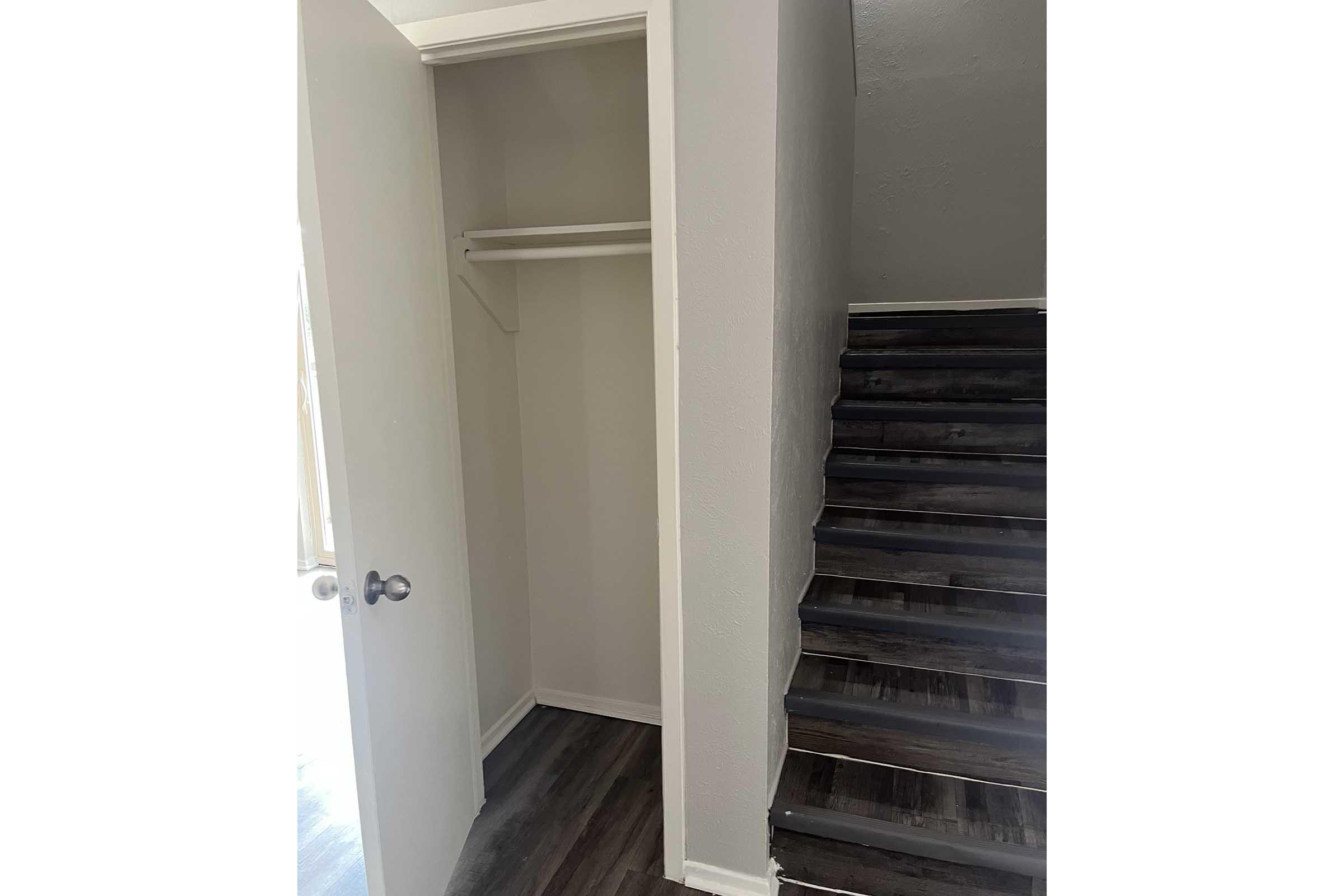 A view of a small closet with a single shelf, featuring a closed door on the left and a staircase with dark wooden steps on the right. The walls are painted light gray, and the floor has a dark wood finish. Natural light is visible, highlighting the space.