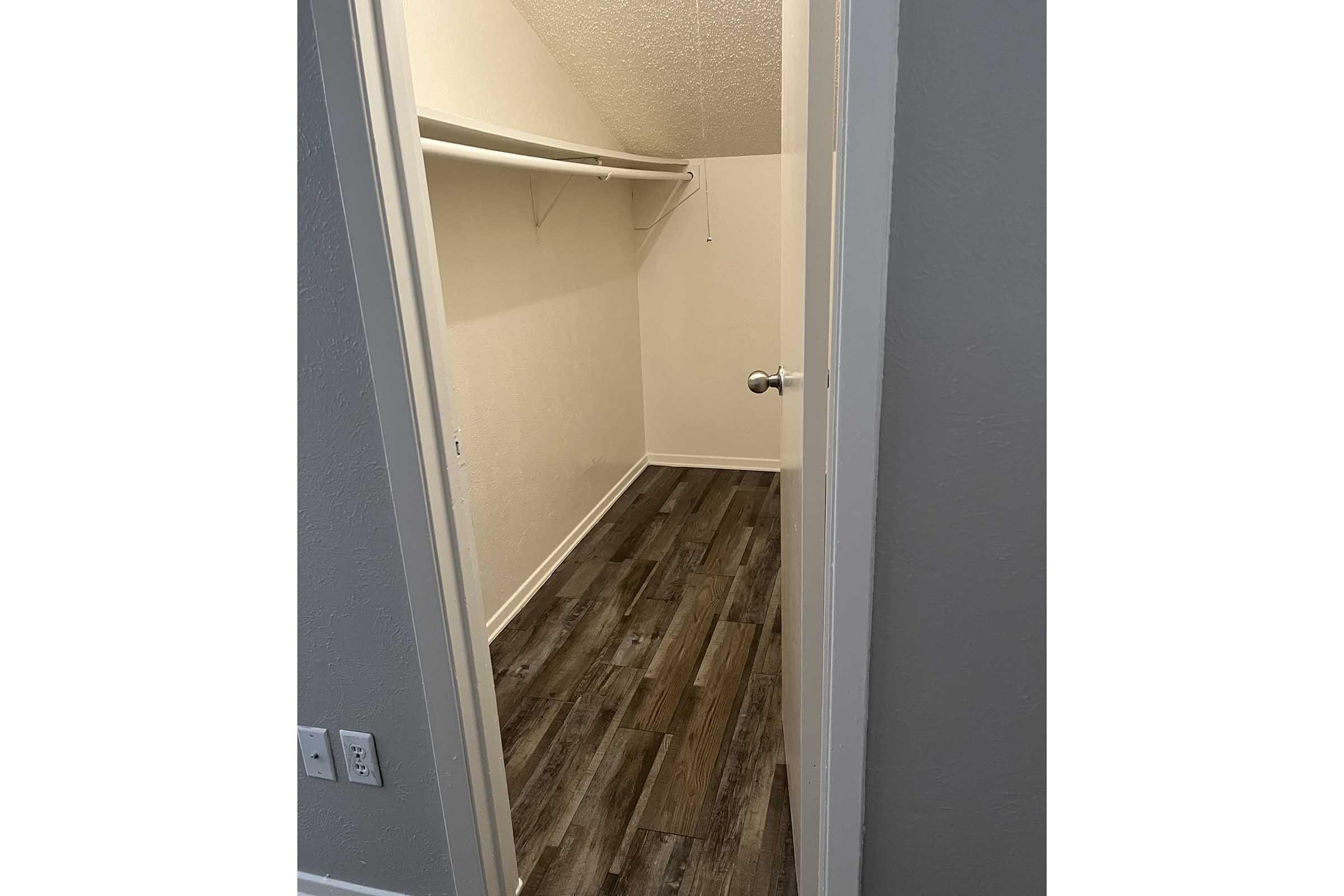 A spacious, empty closet with light-colored walls and dark wood-like flooring. The closet features a single shelf along the back wall, and the door is open, revealing the interior space. There are no items stored inside, creating a clean and organized appearance.