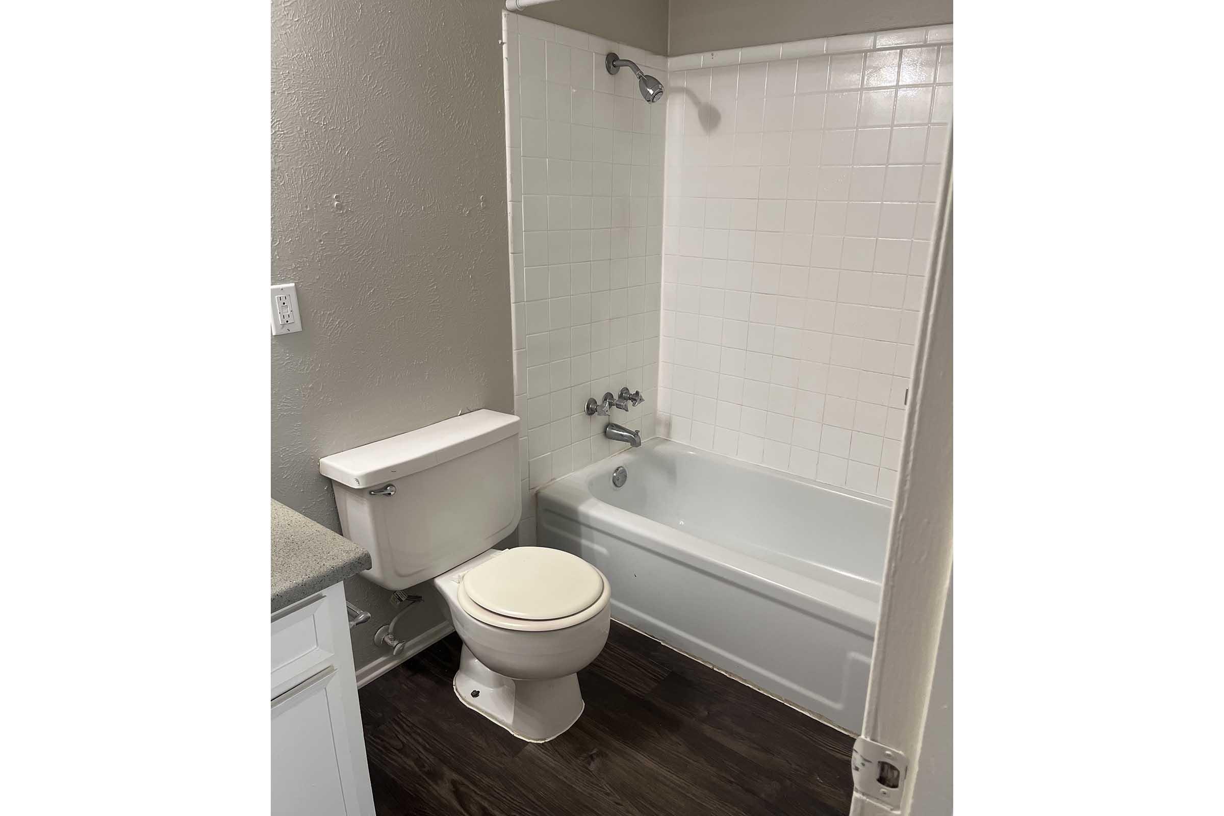 A small bathroom featuring a bathtub and shower combination, a white toilet, and a countertop. The walls are painted a light gray and there are white tiles surrounding the bathtub. The floor has a dark wooden finish, adding contrast to the overall neutral color scheme.