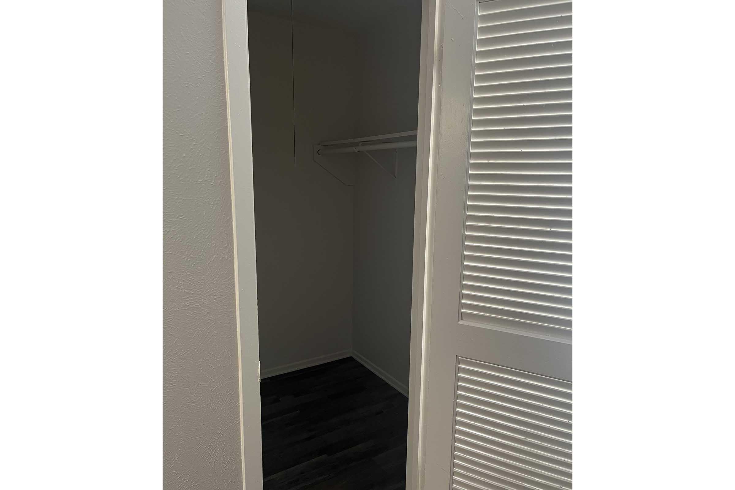 A partially open white door revealing a small, empty closet with a wall shelf. The closet has light-colored walls and dark flooring, creating a simple, clean appearance.