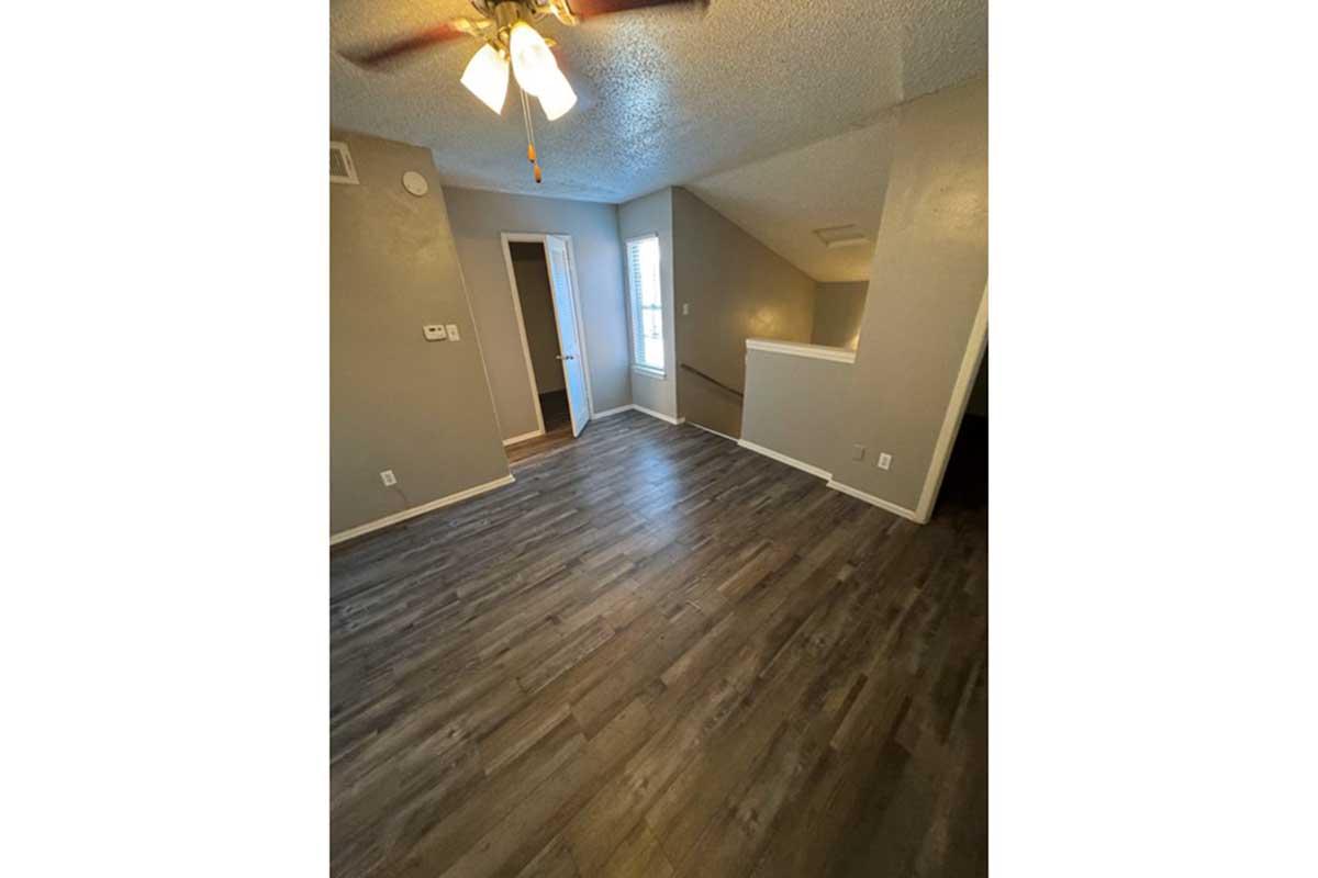 A spacious, well-lit room with grey walls and wooden flooring. A ceiling fan is visible, and there are doors leading to other areas. Natural light filters in through a window, creating a cozy atmosphere. The layout features an open space, ideal for various furniture arrangements.