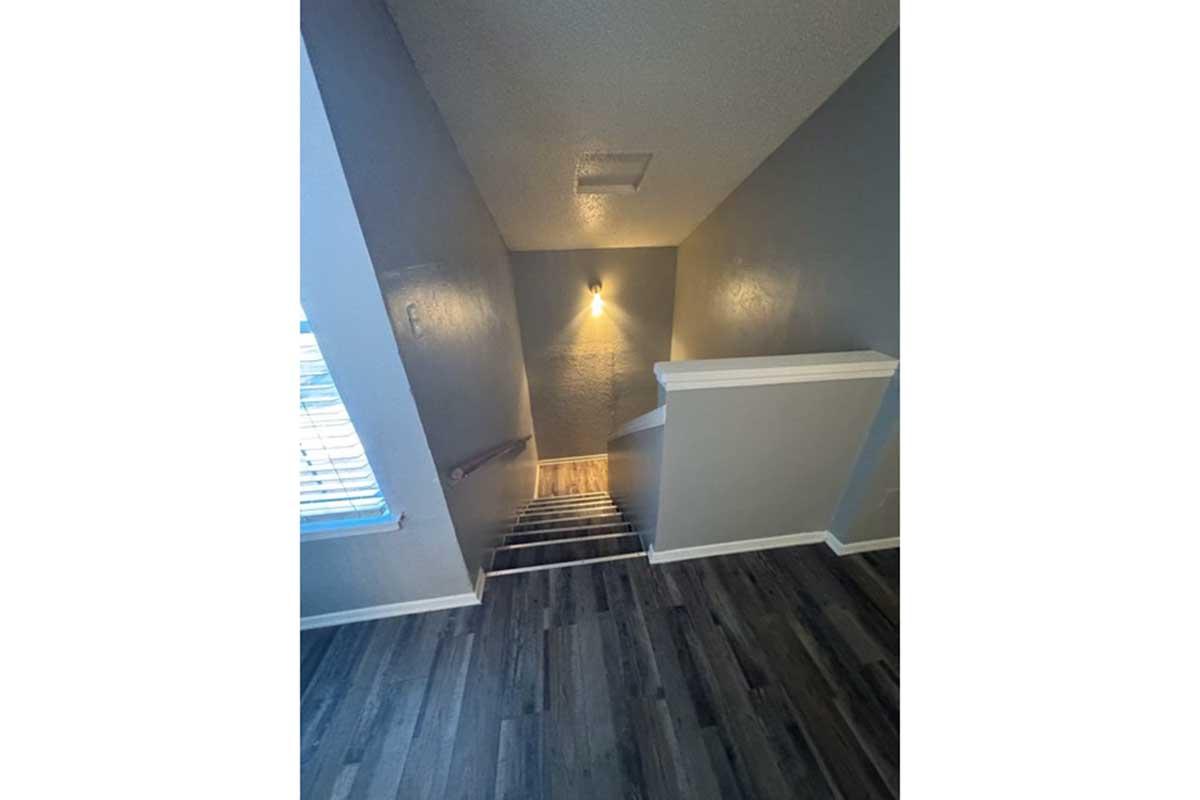 A narrow staircase leading downwards, featuring a light fixture on the wall. The walls are painted in a light gray color, and the floor has wooden planks. A window with blinds is visible at the top of the stairs, allowing natural light to enter the space.