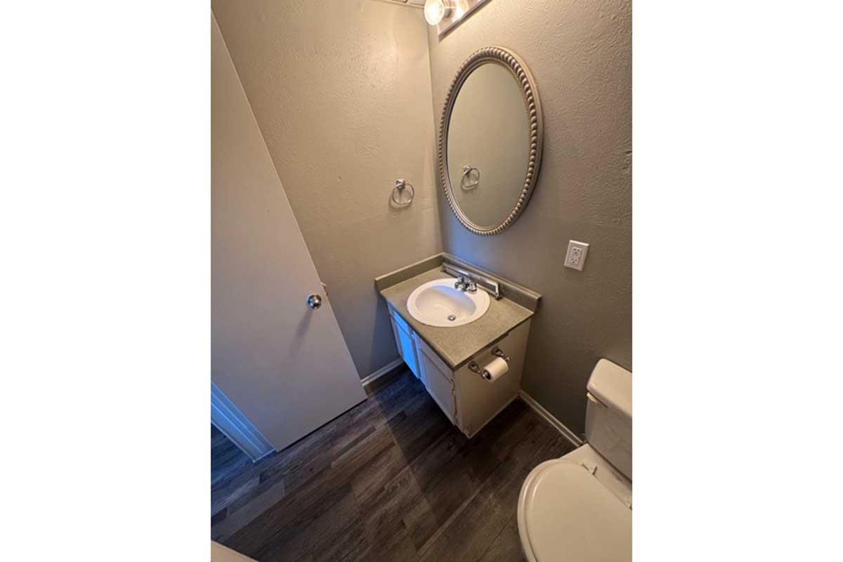 A modern bathroom featuring a small sink with a round mirror above it, a towel holder on the wall, and a toilet nearby. The walls are painted a light color, and the flooring is a wood-like finish, creating a clean and simple design.