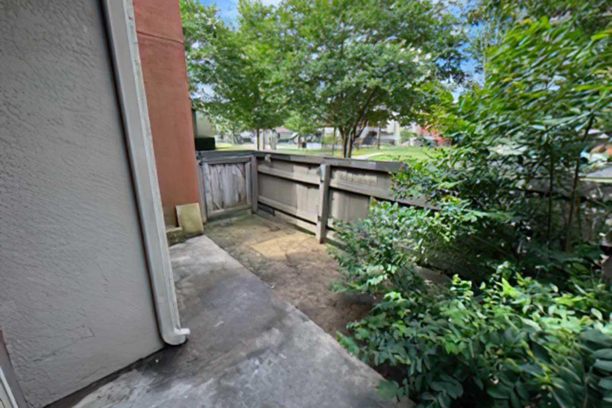 View of a narrow outdoor walkway between a building and a wooden fence, lined with bushes and greenery. The area is well-lit by natural sunlight, with trees visible in the background and a gravel path leading into the space.