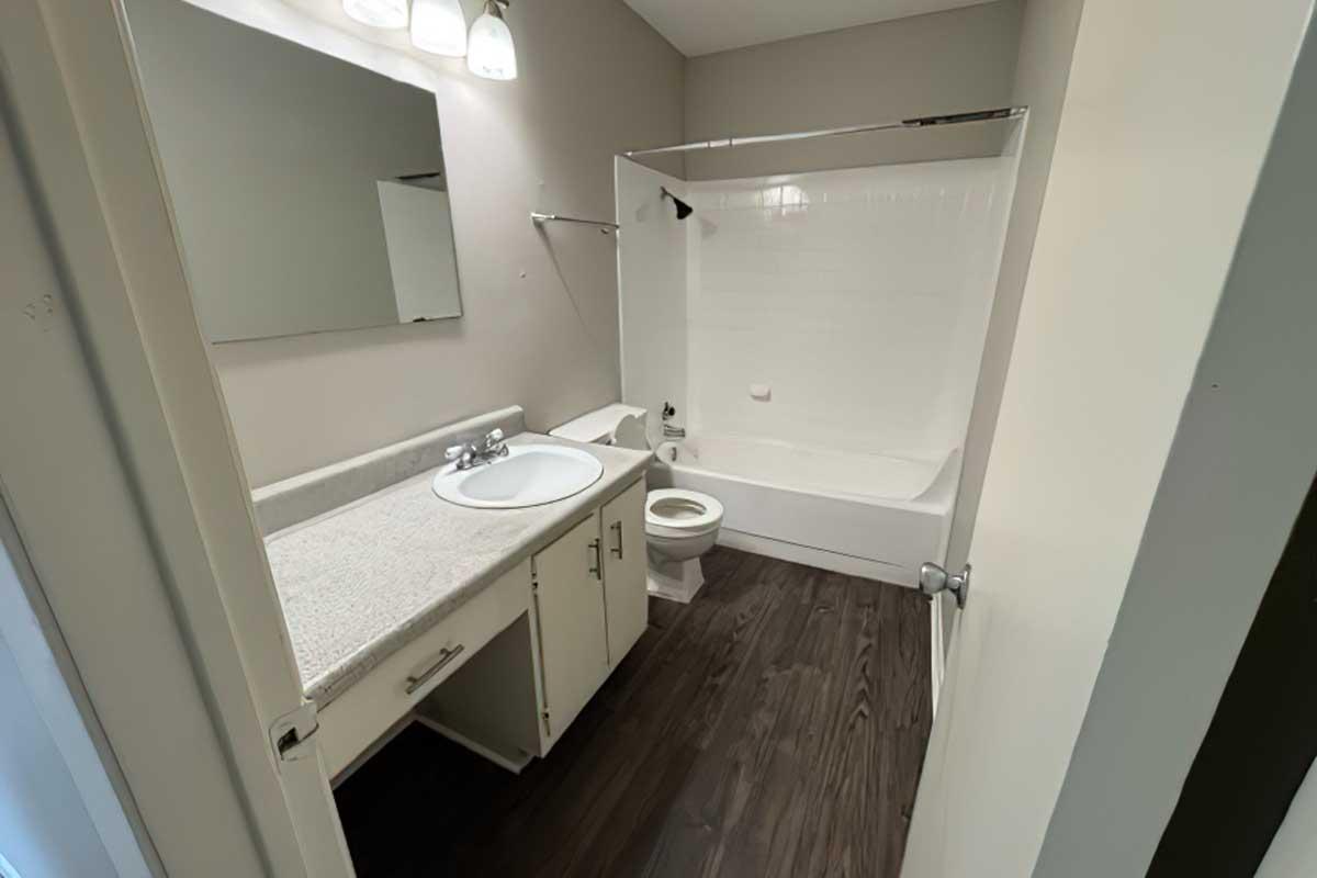 A clean, modern bathroom featuring a white bathtub with a shower, a vanity with a sink, and a toilet. The walls are a light color, and there is a rectangular mirror above the sink. The flooring is dark, adding contrast to the overall decor.